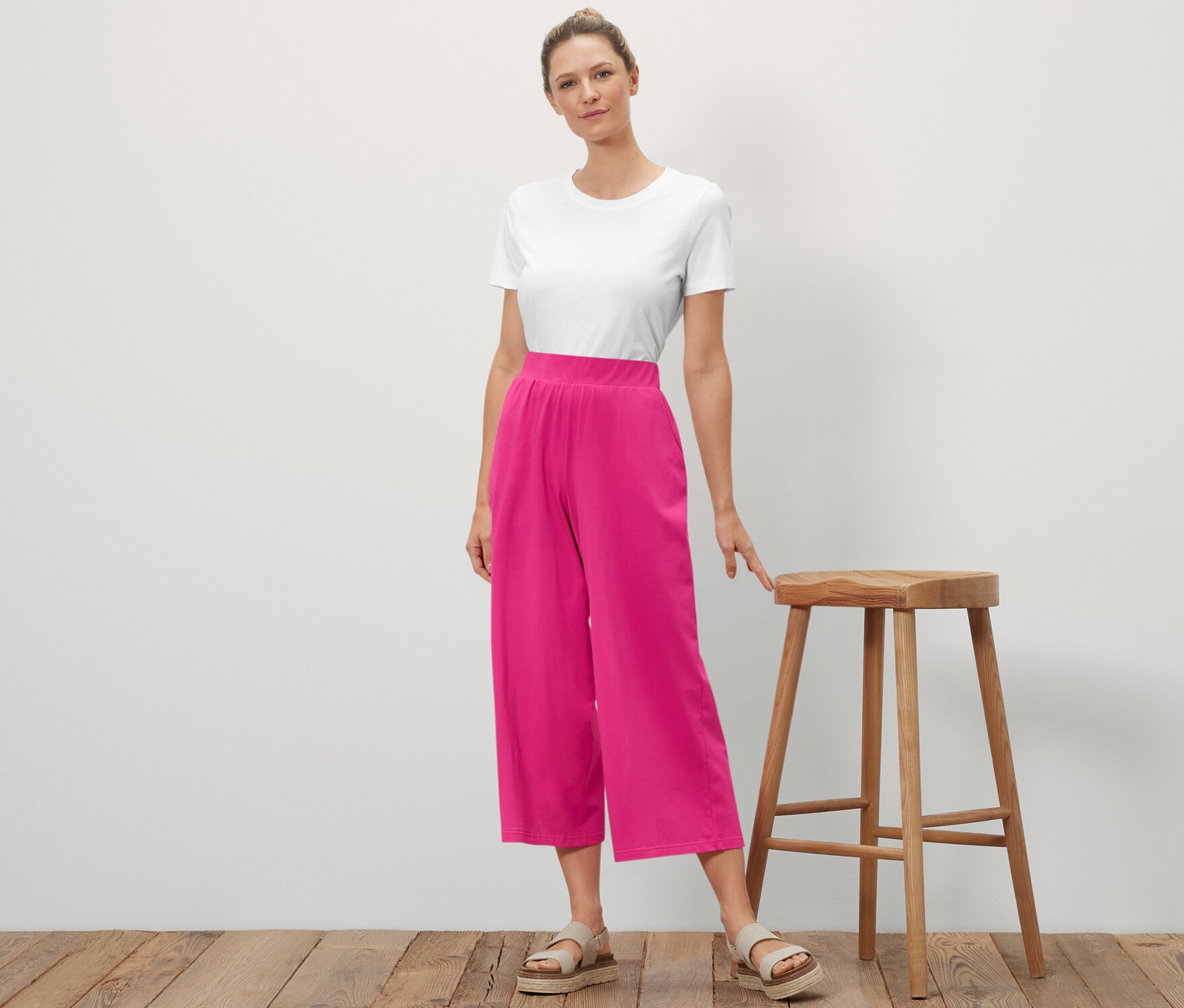 Kobieta stoi i pozuje obok drewnianego stołka. Ma na sobie biały t-shirt i różowe spodnie kuloty z dżerseju.