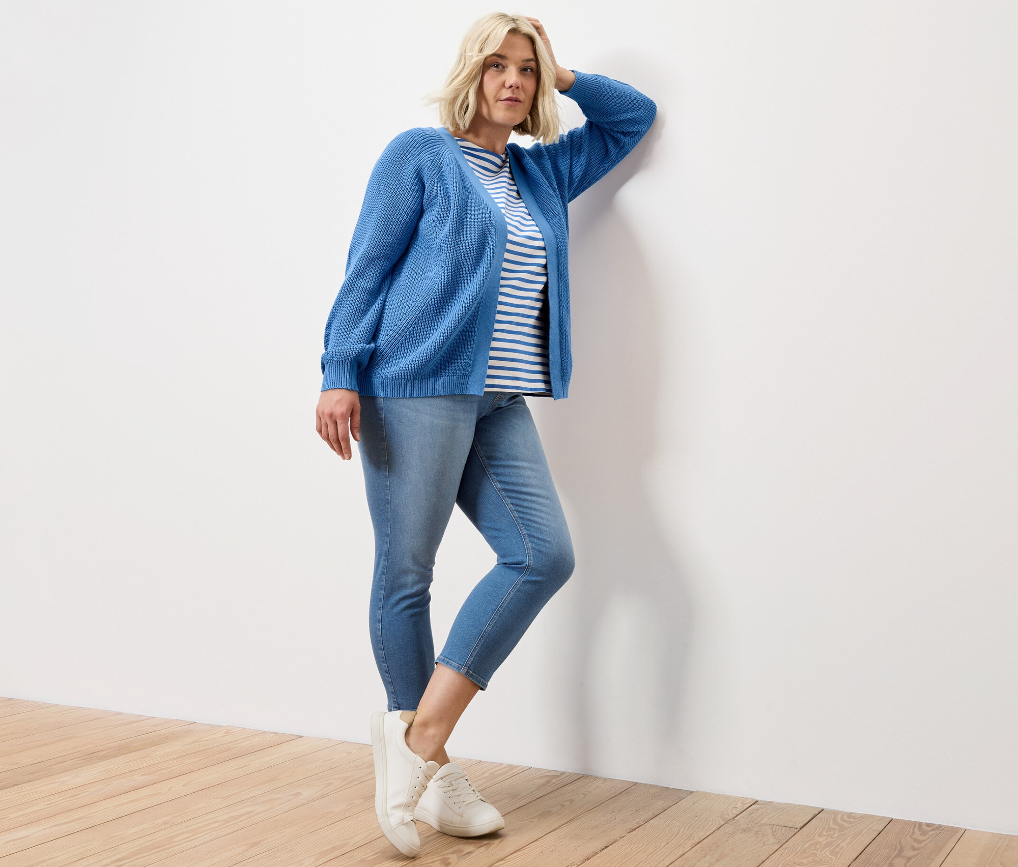 Kobieta pozuje oparta o ścianę w koszulce w niebieskie paski z półdługim rękawem, tregginsach 7/8 – fason „Nelly” i kardiganie.