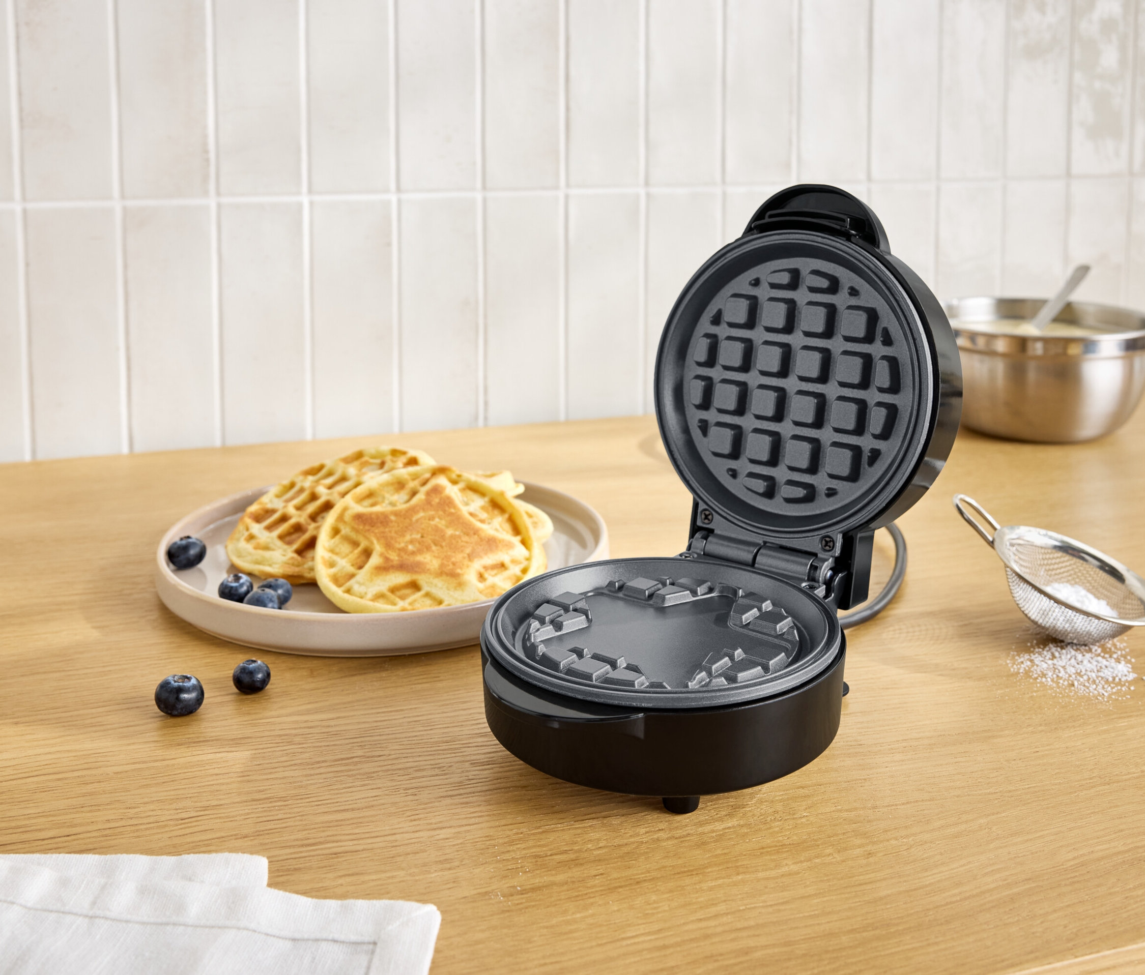 Otwarty, czarny gofrownica obok talerza z goframi i jagodami na drewnianym stole.