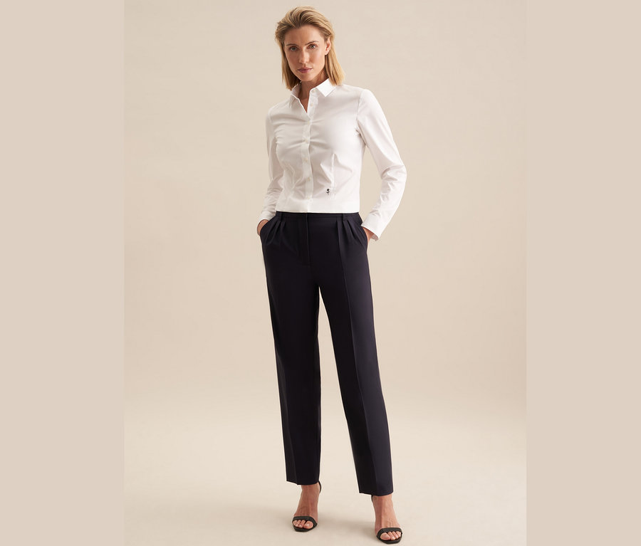Kobieta stoi z rękami w kieszeniach spodni. Ma na sobie białą koszulę slim fit Seidensticker, granatowe spodnie i czarne sandały.