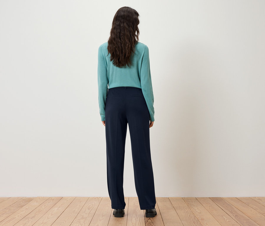 Widok z tyłu kobiety z ciemnobrązowymi włosami w turkusowym swetrze i granatowych spodniach.