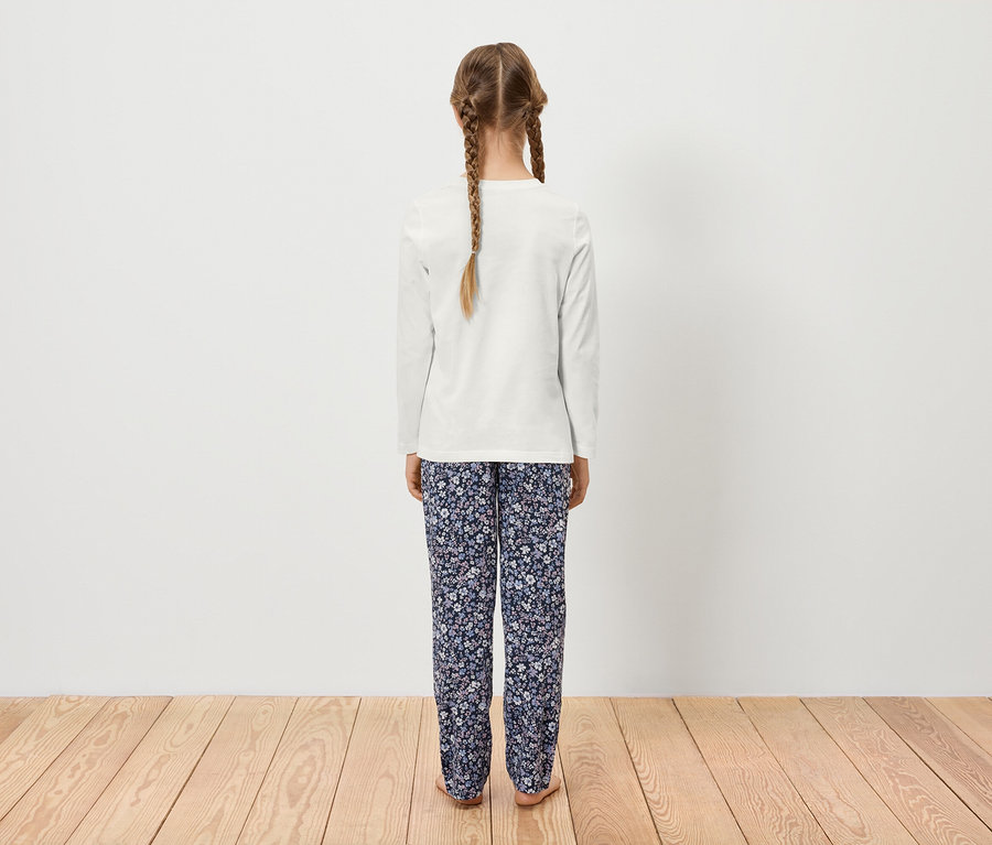 Dziewczyna z dwoma warkoczami stoi tyłem do kamery, ma na sobie białą bluzkę z długim rękawem i kwieciste spodnie.