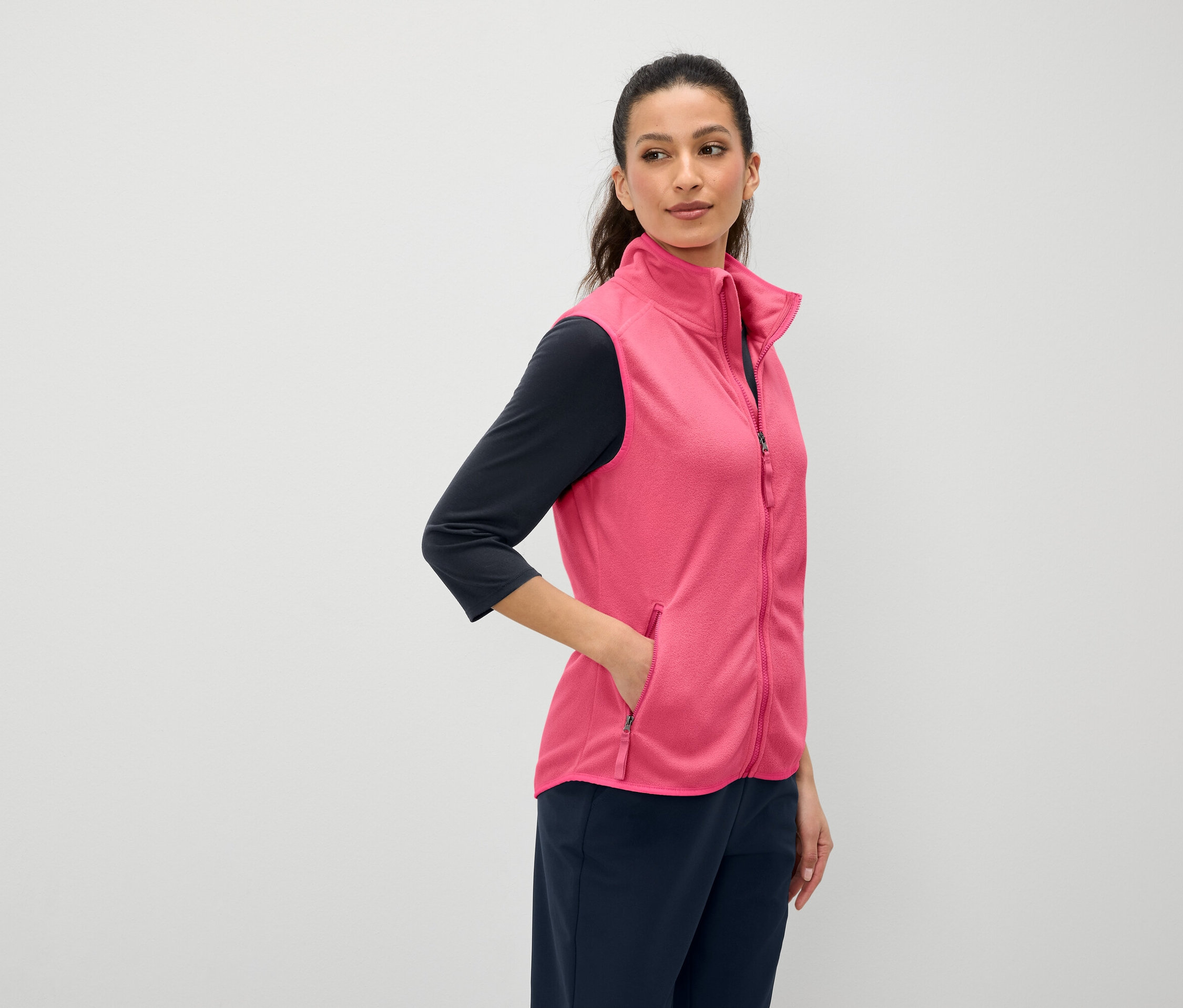 Kobieta o brązowych włosach trzyma rękę w kieszeni różowej kamizelki. Ma na sobie granatową koszulkę z rękawem trzy czwarte i granatowe spodnie.