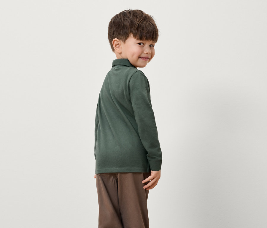 Chłopiec spogląda przez ramię, ubrany w zieloną koszulkę polo z długim rękawem i brązowe spodnie.