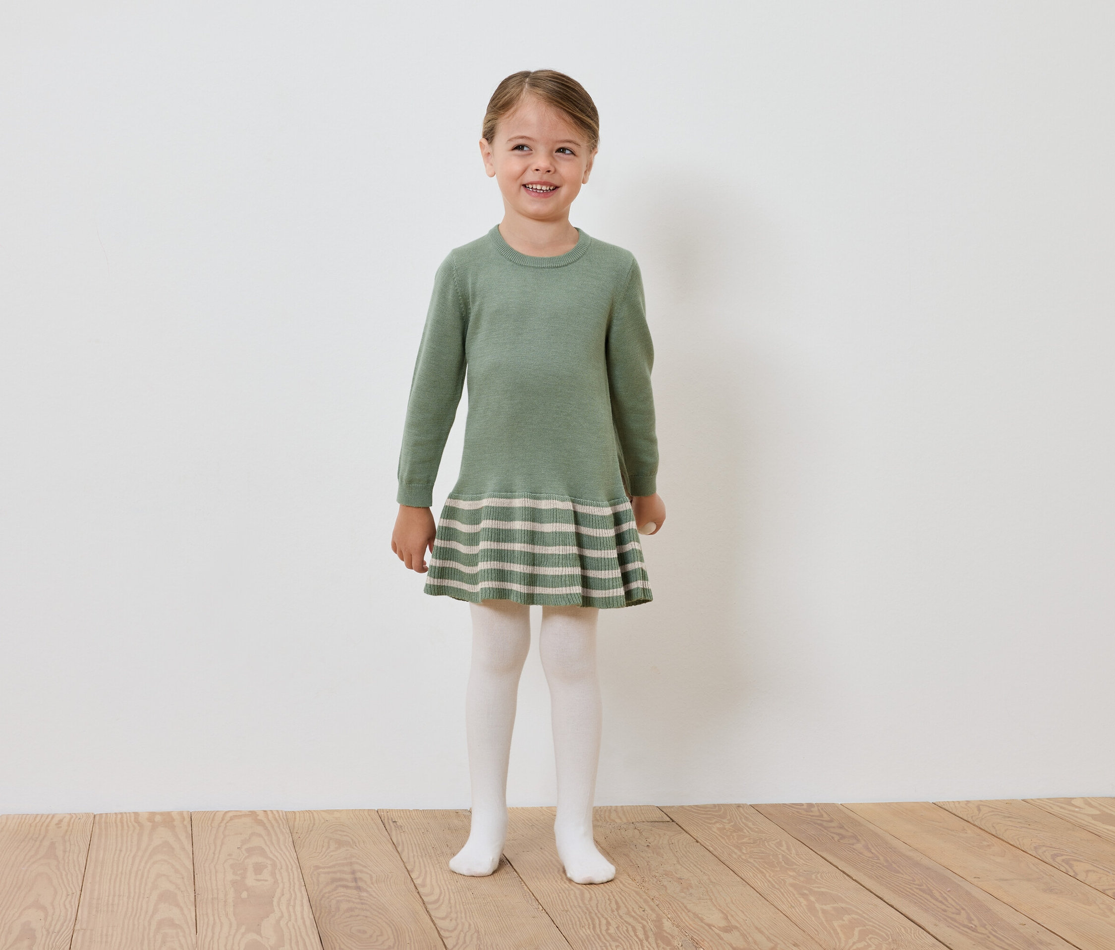 Uśmiechnięta dziewczynka w zielonej dzianinowej sukience i białych rajstopach stoi na drewnianej podłodze przed białą ścianą.