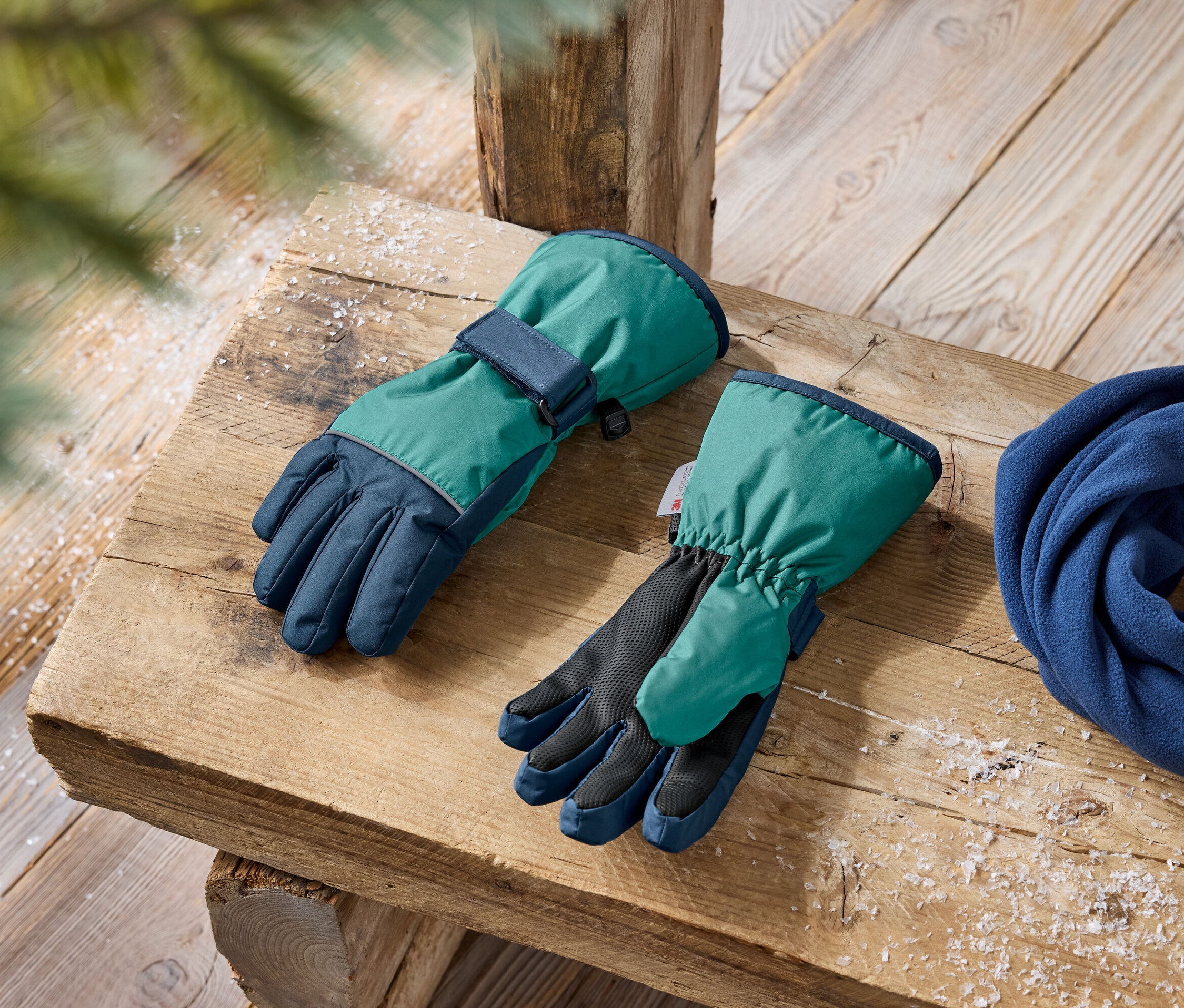 Zielono-granatowe rękawice zimowe i niebieski polarowy szal leżą na drewnianej ławce.