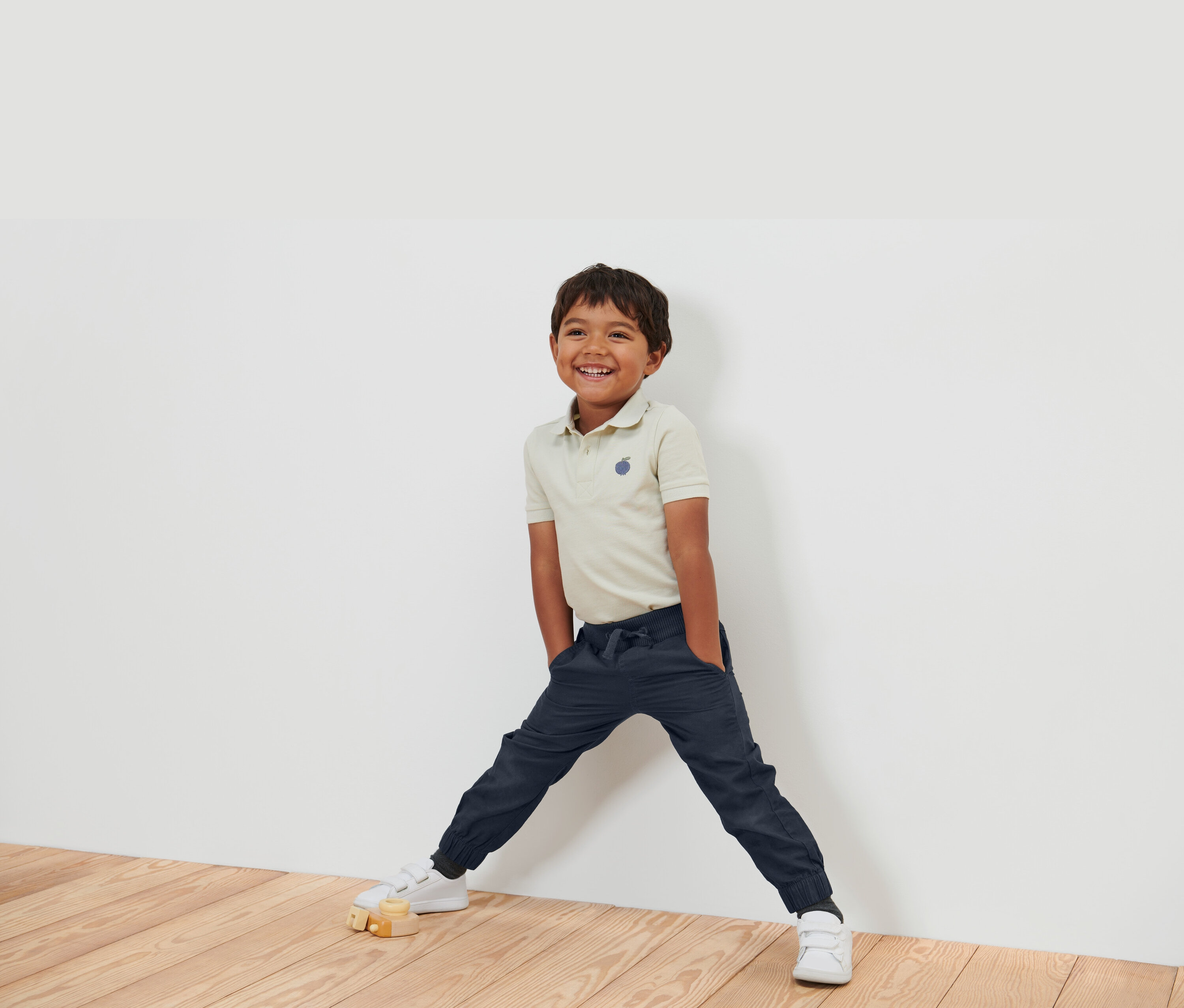 Uśmiechnięty chłopiec w beżowej koszulce polo, granatowych spodniach i białych butach stoi oparty o białą ścianę.