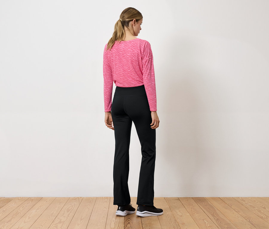 Kobieta tyłem do kamery ma na sobie różową bluzkę z długim rękawem i czarne spodnie.