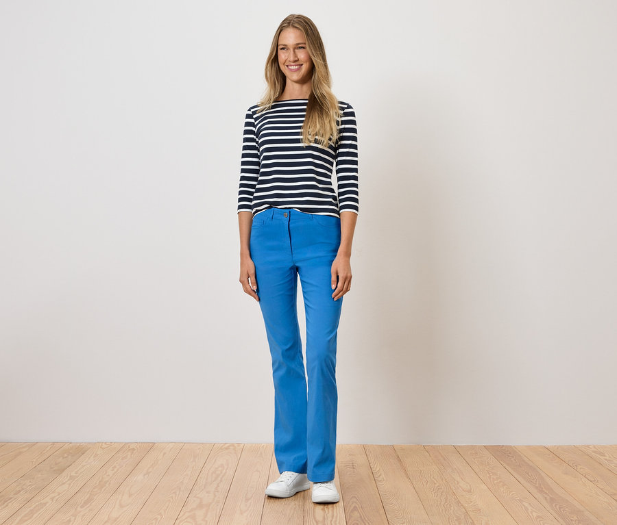 Kobieta stoi, ubrana w niebieskie spodnie, koszulkę w paski i białe buty.