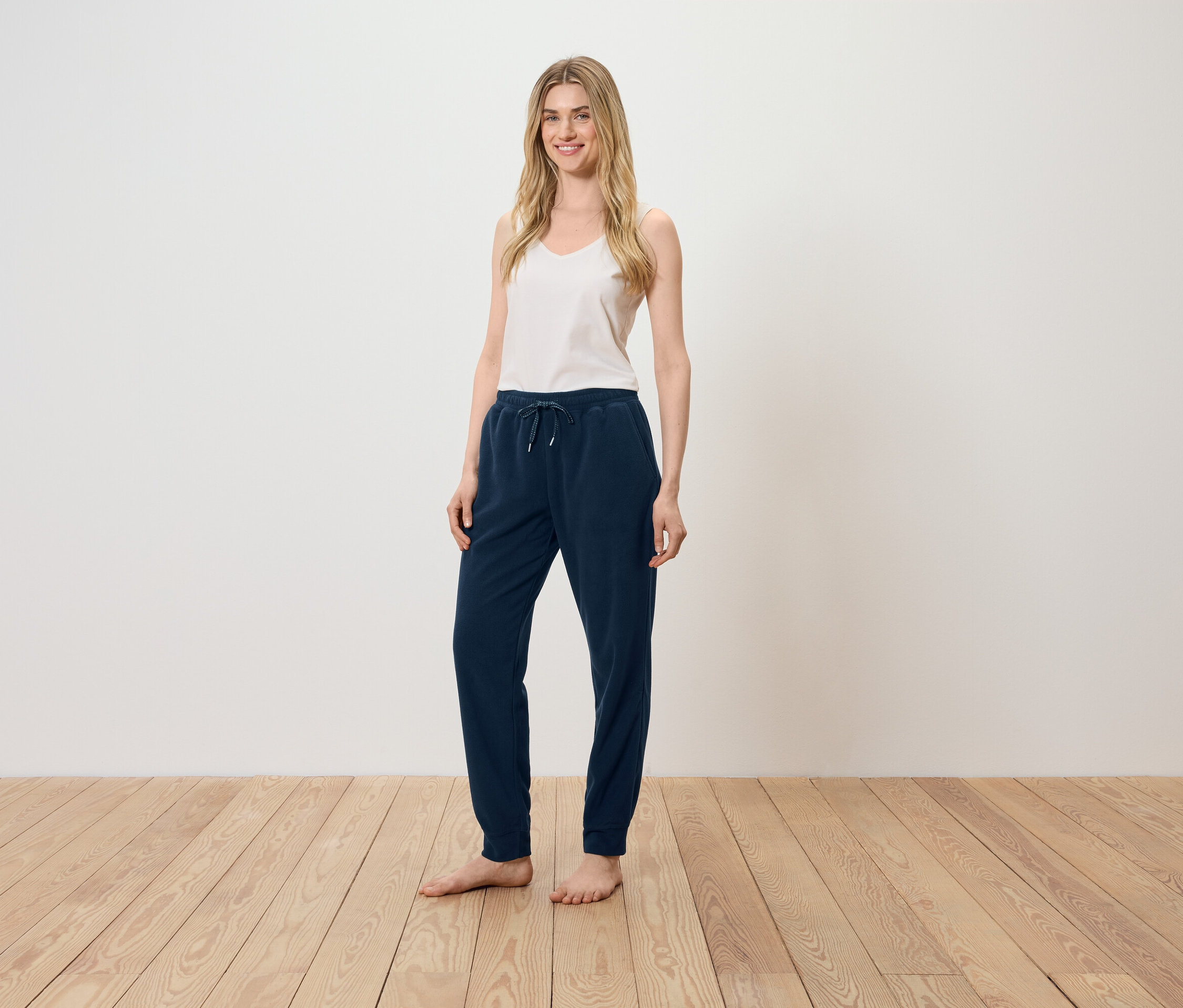 Kobieta stoi na drewnianej podłodze, ubrana w biały top i granatowe spodnie dresowe.