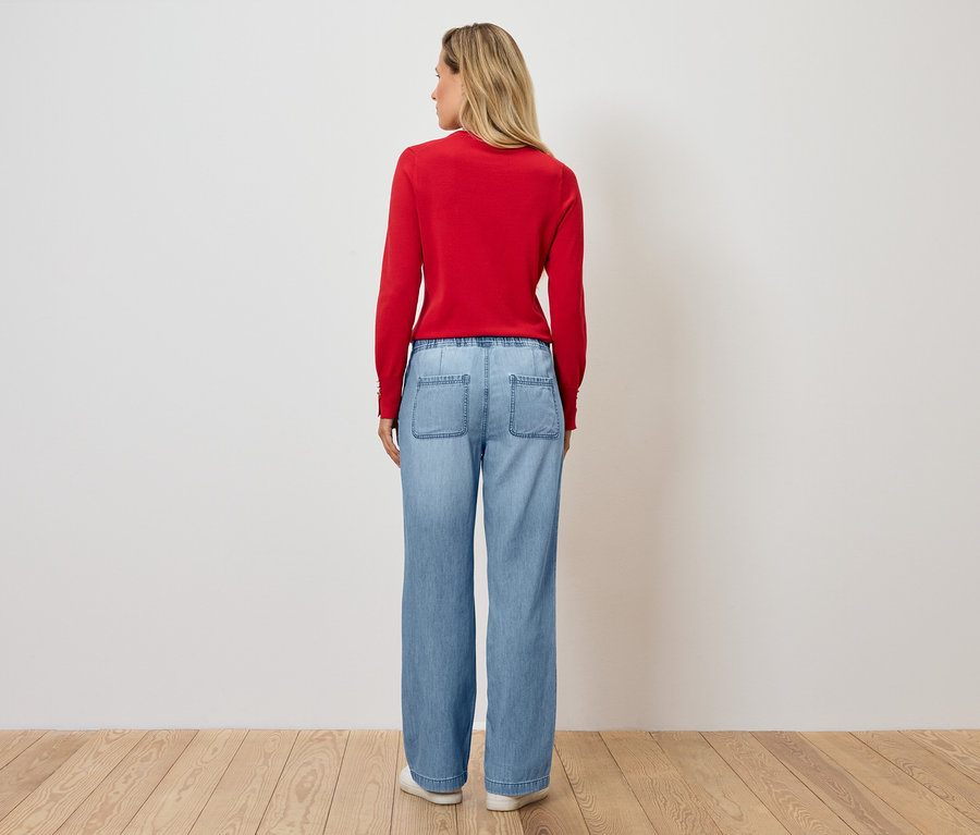 Kobieta stoi tyłem do aparatu, ubrana w czerwony sweter i niebieskie dżinsy.