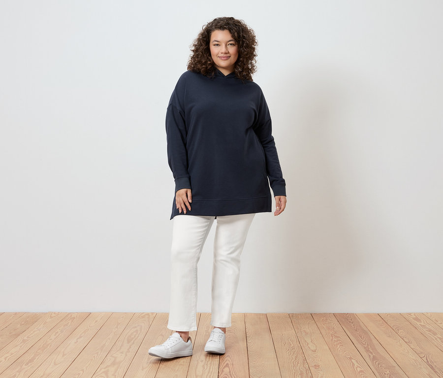 Kobieta o kręconych włosach stoi na tle ściany, ubrana w granatową bluzę z kapturem, białe dżinsy i białe tenisówki.