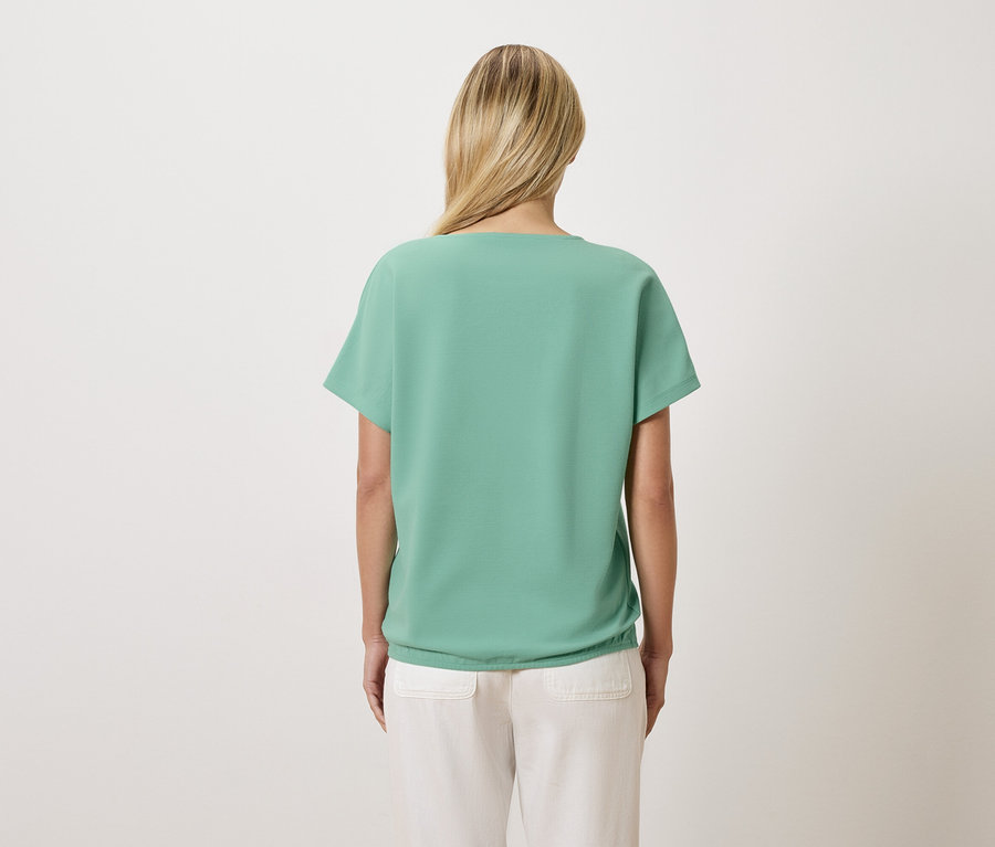 Widok z tyłu kobiety o blond włosach, ubranej w jasnozielony T-shirt i jasne spodnie.