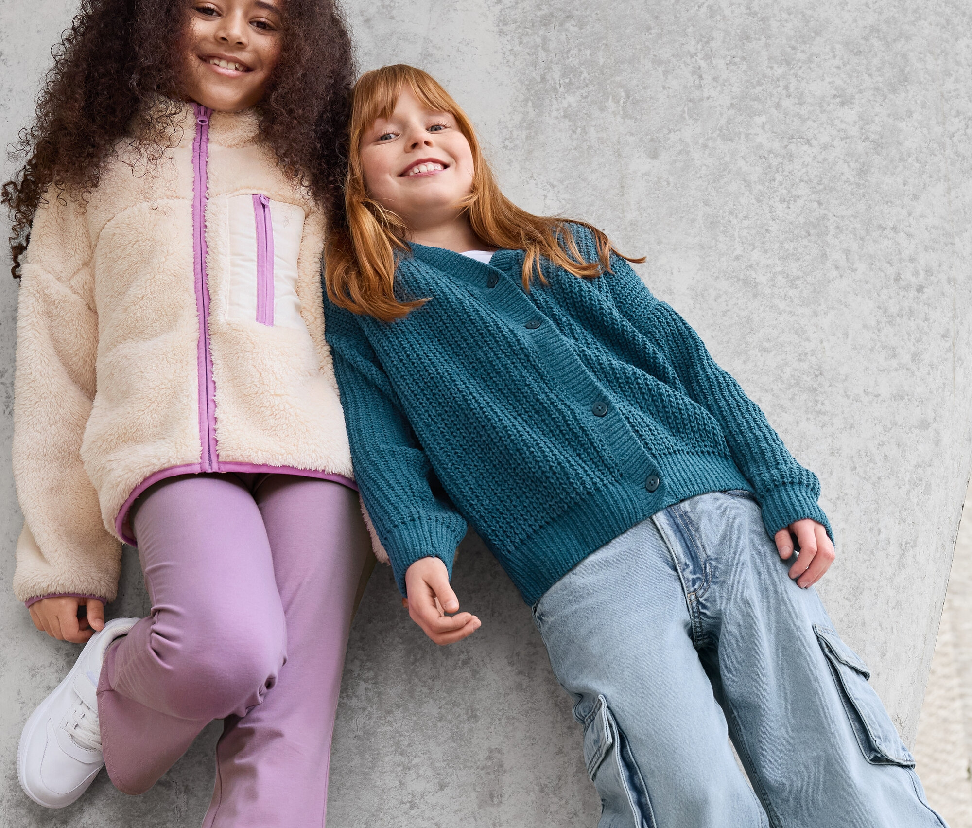 Dwie dziewczynki opierają się o betonową ścianę. Dziewczynka po lewej ma na sobie kurtkę z polaru, fioletowe spodnie i białe tenisówki. Dziewczynka po prawej ma na sobie zielony kardigan i niebieskie spodnie.