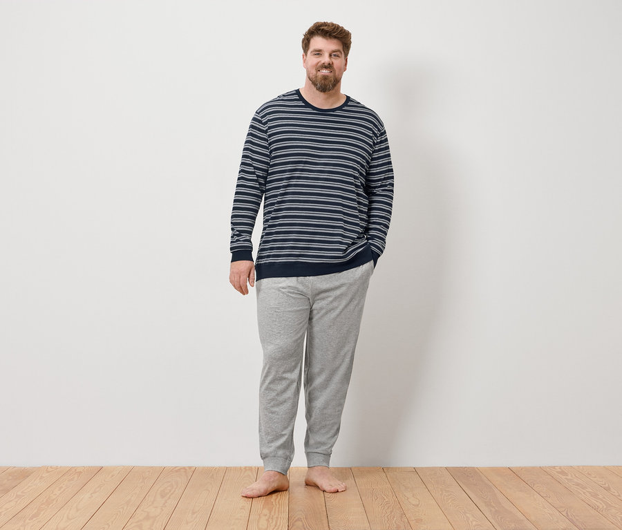 Mężczyzna stoi boso przed ścianą, ubrany w szarą piżamę.