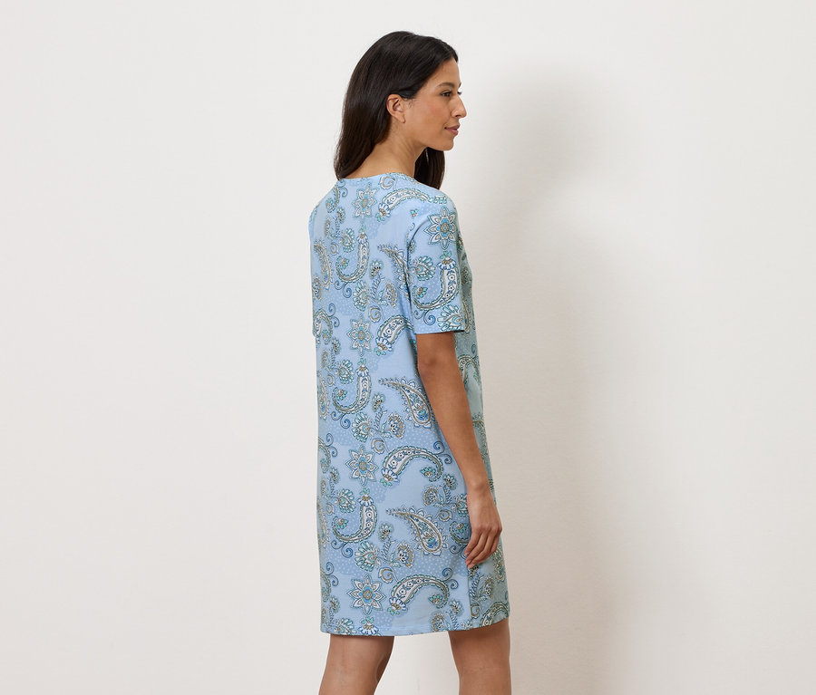 Kobieta stoi bokiem, ubrana w jasnoniebieską sukienkę w kwiaty.
