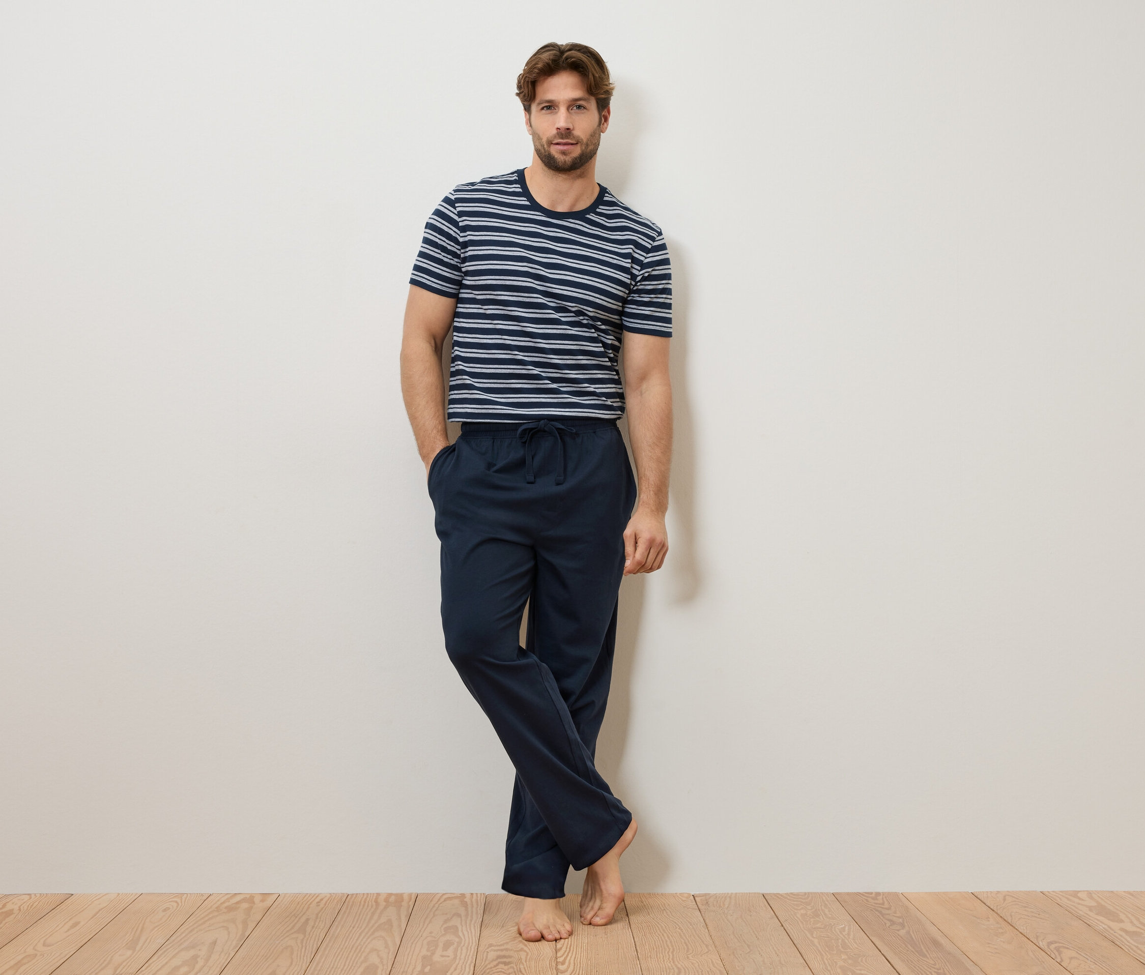 Mężczyzna opiera się o ścianę, ubrany w granatowo-biały T-shirt w paski i granatowe spodnie od piżamy.