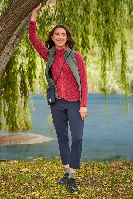 Kobieta opiera się o drzewo nad jeziorem, ubrana w czerwoną bluzę polarową, granatowe spodnie i khaki kamizelkę »packable« Hybrid-Weste.