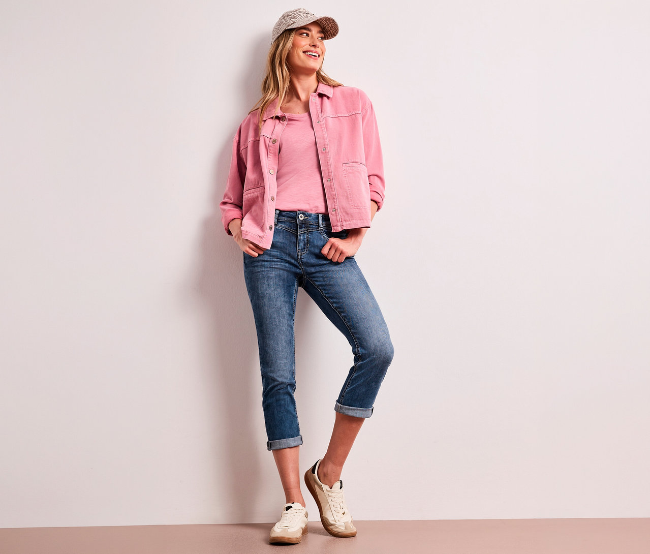 Uśmiechnięta kobieta w czapce z daszkiem, różowej kurtce, różowym T-shircie i niebieskich jeansach 7/8 Street One „Jane” z podwiniętymi nogawkami, oparta o białą ścianę.