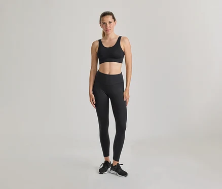 Kobieta stoi, ubrana w czarny biustonosz sportowy i czarne legginsy. Czarne i białe sneakersy, włosy związane. Jasne, jednolite tło.