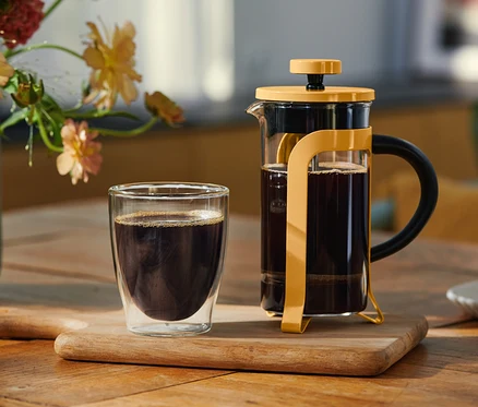 Szklanka czarnej kawy z podwójnymi ściankami i żółty zaparzacz French Press wypełniony kawą stoją na drewnianej desce. W tle wazon z kwiatami i talerzyk z ciastkiem.
