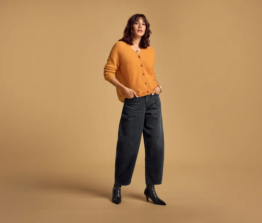 Kobieta w pomarańczowym Merino-Cardiganie i czarnych Barrel Jeans, z rękami w kieszeniach.