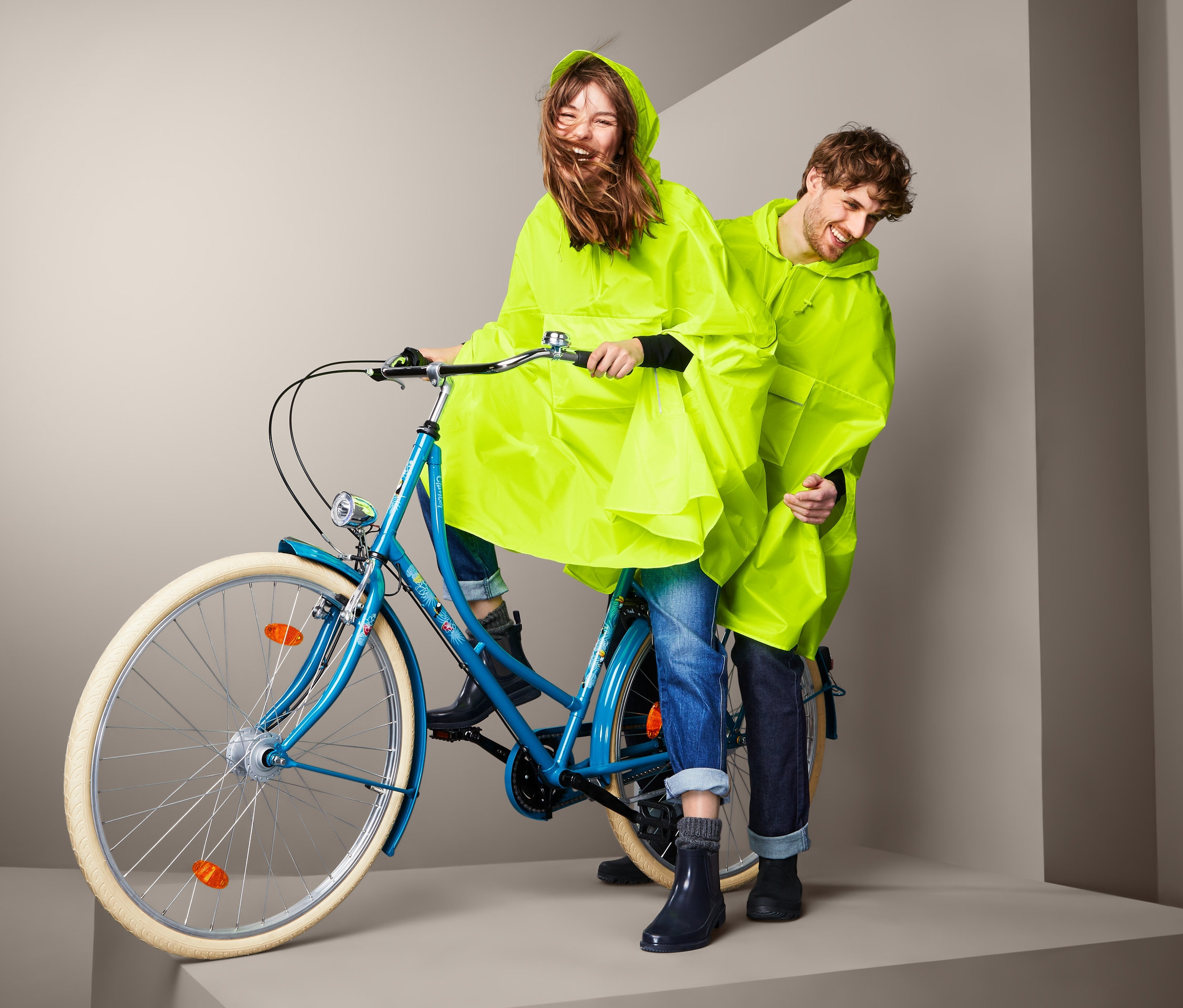 Kobieta jedzie na niebieskim rowerze z mężczyzną z tyłu, oboje w żółtych ponczach przeciwdeszczowych.