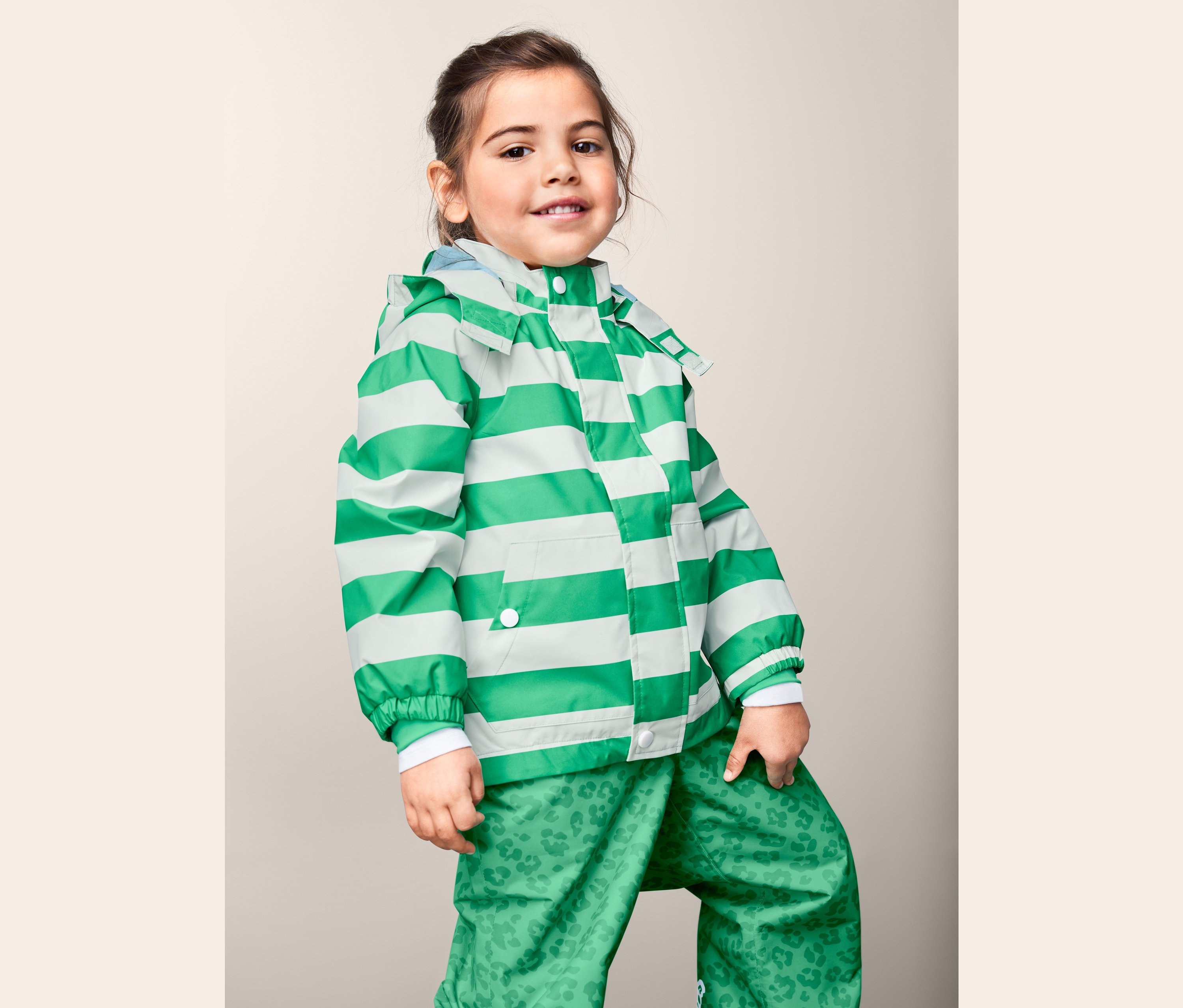 Dziewczynka pozuje w kurtce przeciwdeszczowej z materiału z recyklingu w zielono-białe paski i zielonych spodniach we wzory.