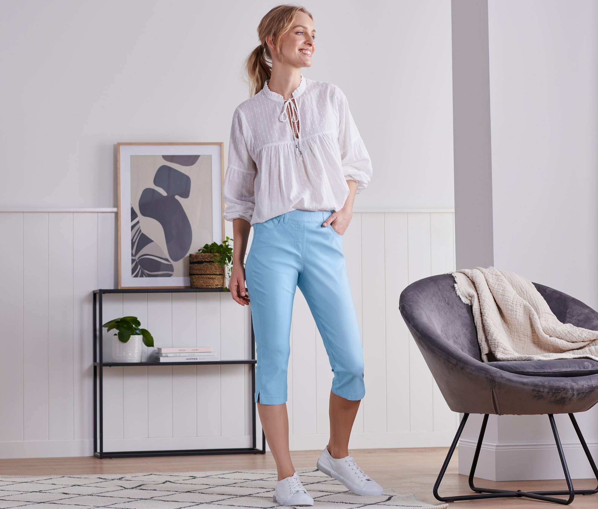 Kobieta stoi w pokoju, ubrana w błękitne spodnie z bengaliny o długości 3/4 i białą bluzkę.