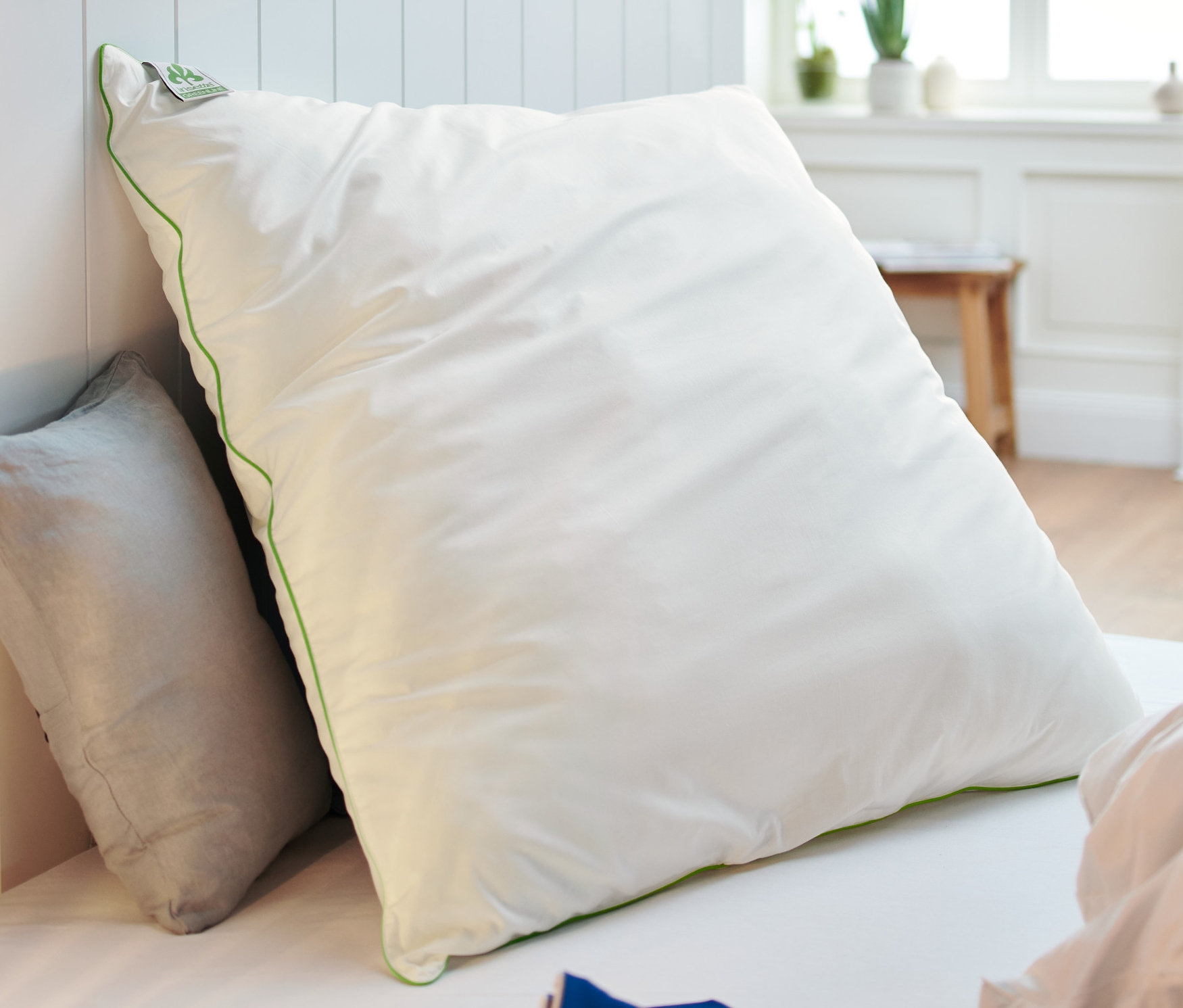 Biała poduszka irisette® greenline o wymiarach ok. 80 x 80 cm oparta o ścianę obok szarej poduszki na łóżku. Na pierwszym planie wiklinowa lampa.