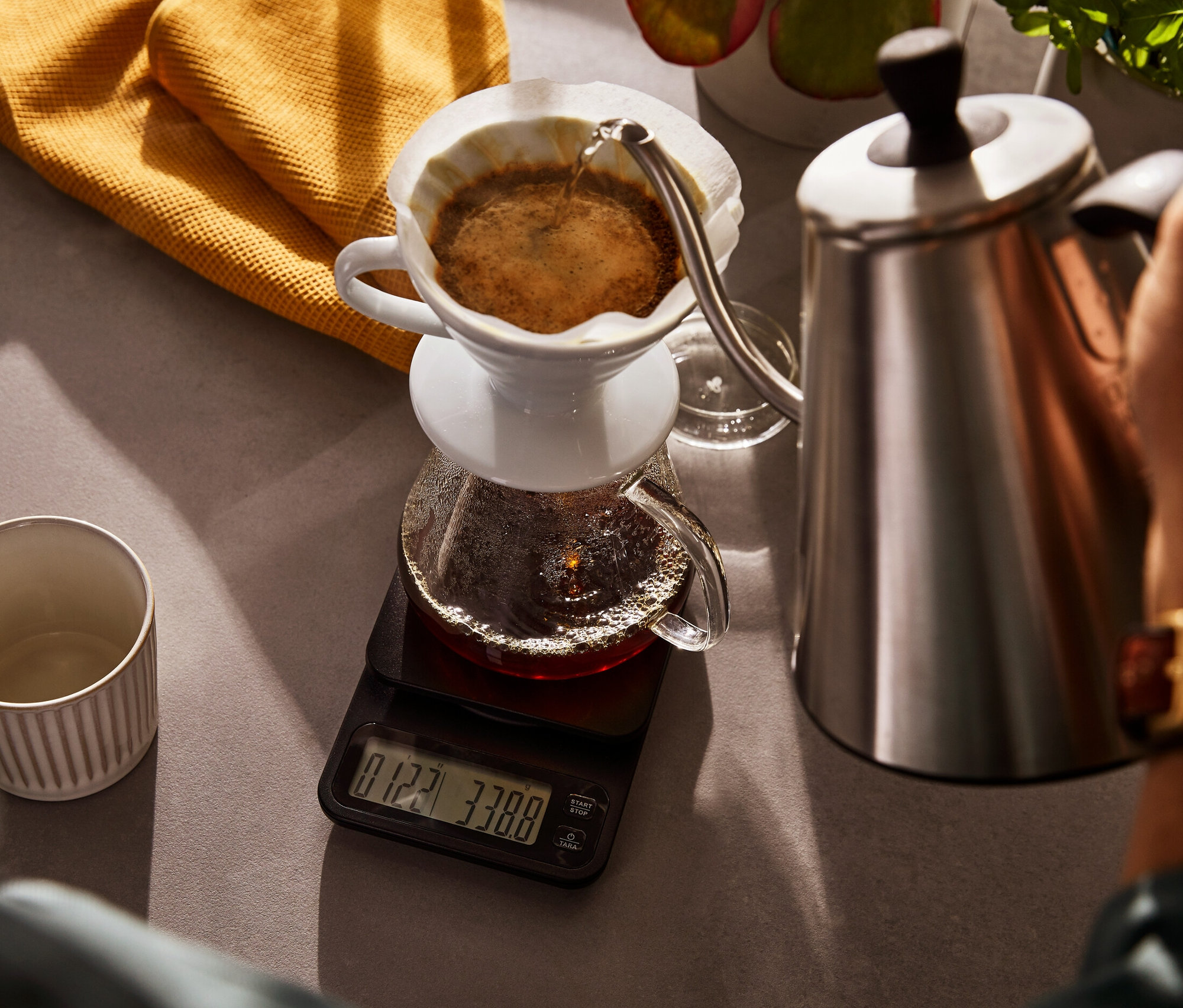 Osoba nalewa wodę z elektrycznego czajnika typu konewka do szklanego dzbanka do kawy stojącego na wadze do kawy z funkcją stopera.