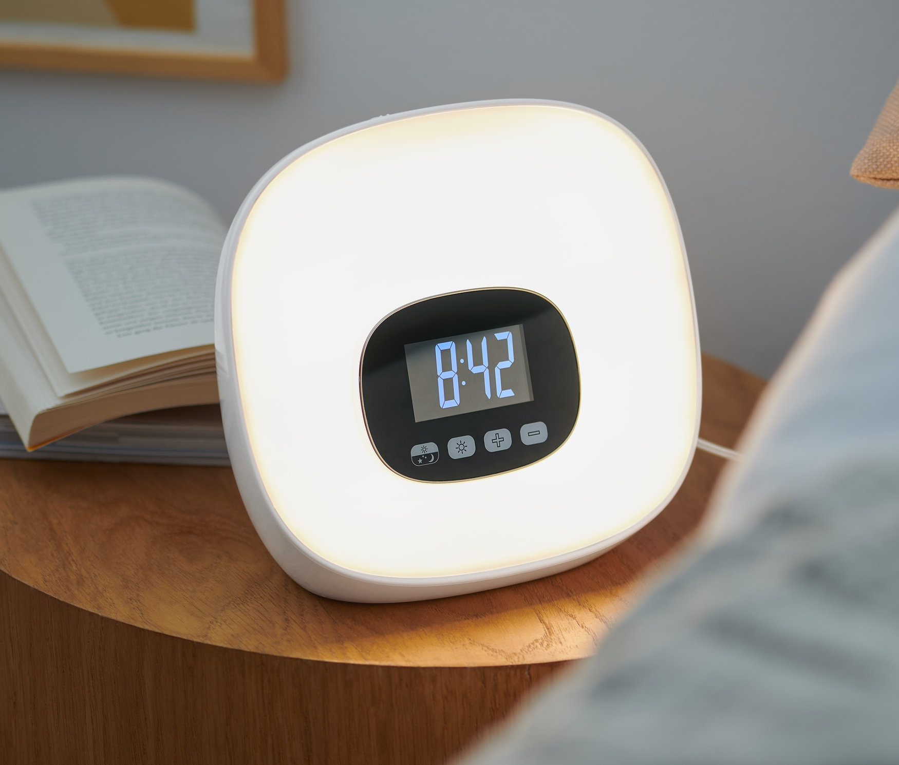 Budzik świetlny z radiem stoi na stoliku nocnym obok książek. Na wyświetlaczu budzika widnieje godzina 8:42.
