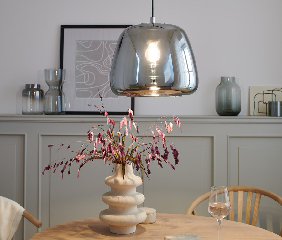 Chromowana lampa wisząca nad stołem z wazonem i kieliszkiem wina.