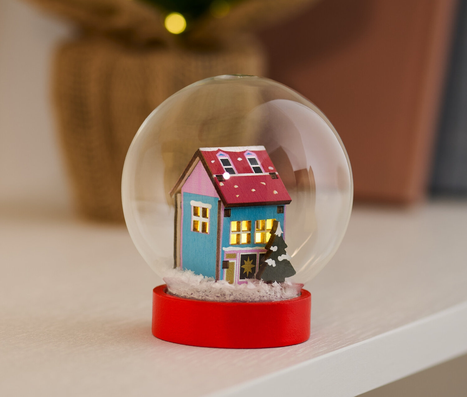 Kula śnieżna LED z niebieskim domkiem i czerwoną podstawą stoi na półce.