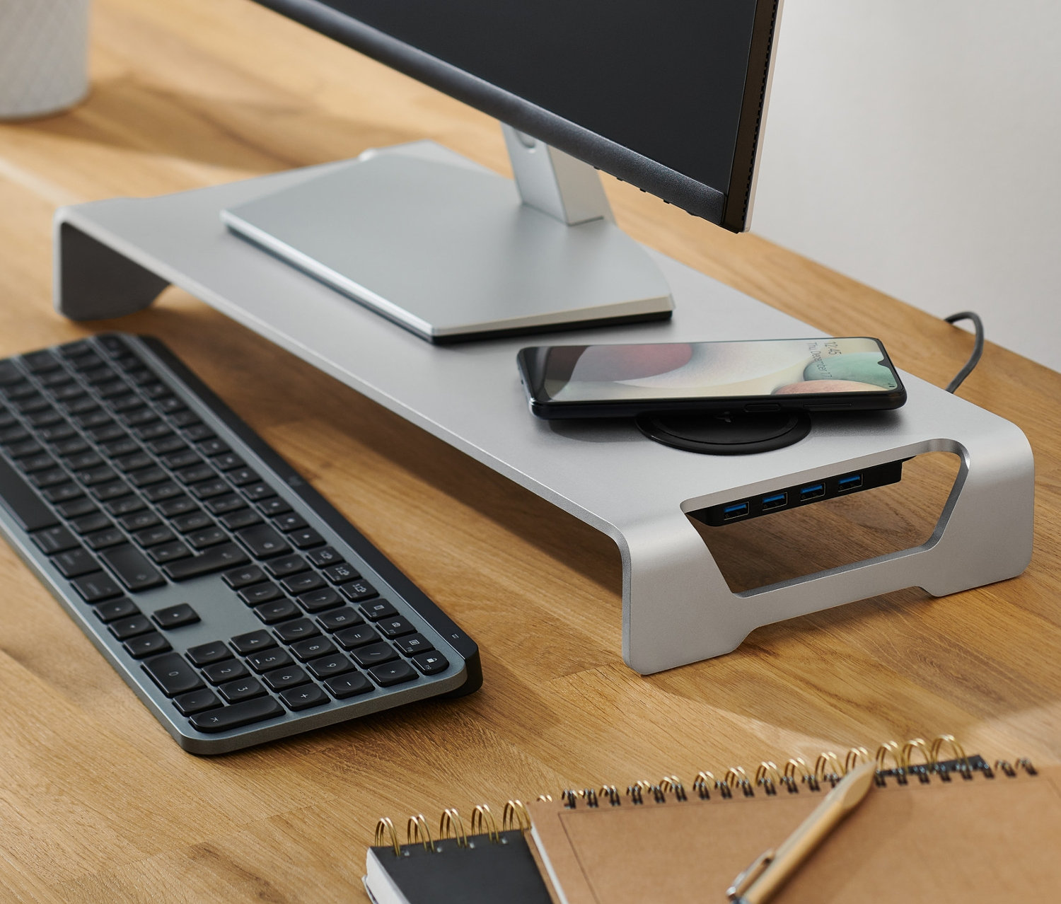 Monitor stoi na srebrnej podstawce pod monitor z bezprzewodową stacją ładowania i hubem USB. Na podstawce leży telefon komórkowy. Obok stoi lampa.