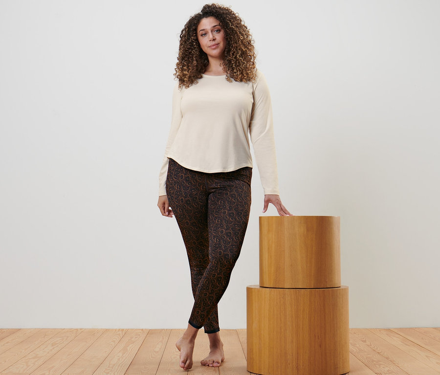 Kobieta pozuje w sportowej koszulce z długim rękawem w kolorze złamanej bieli i dwustronnych legginsach sportowych w kolorze czarnym i brązowym.
