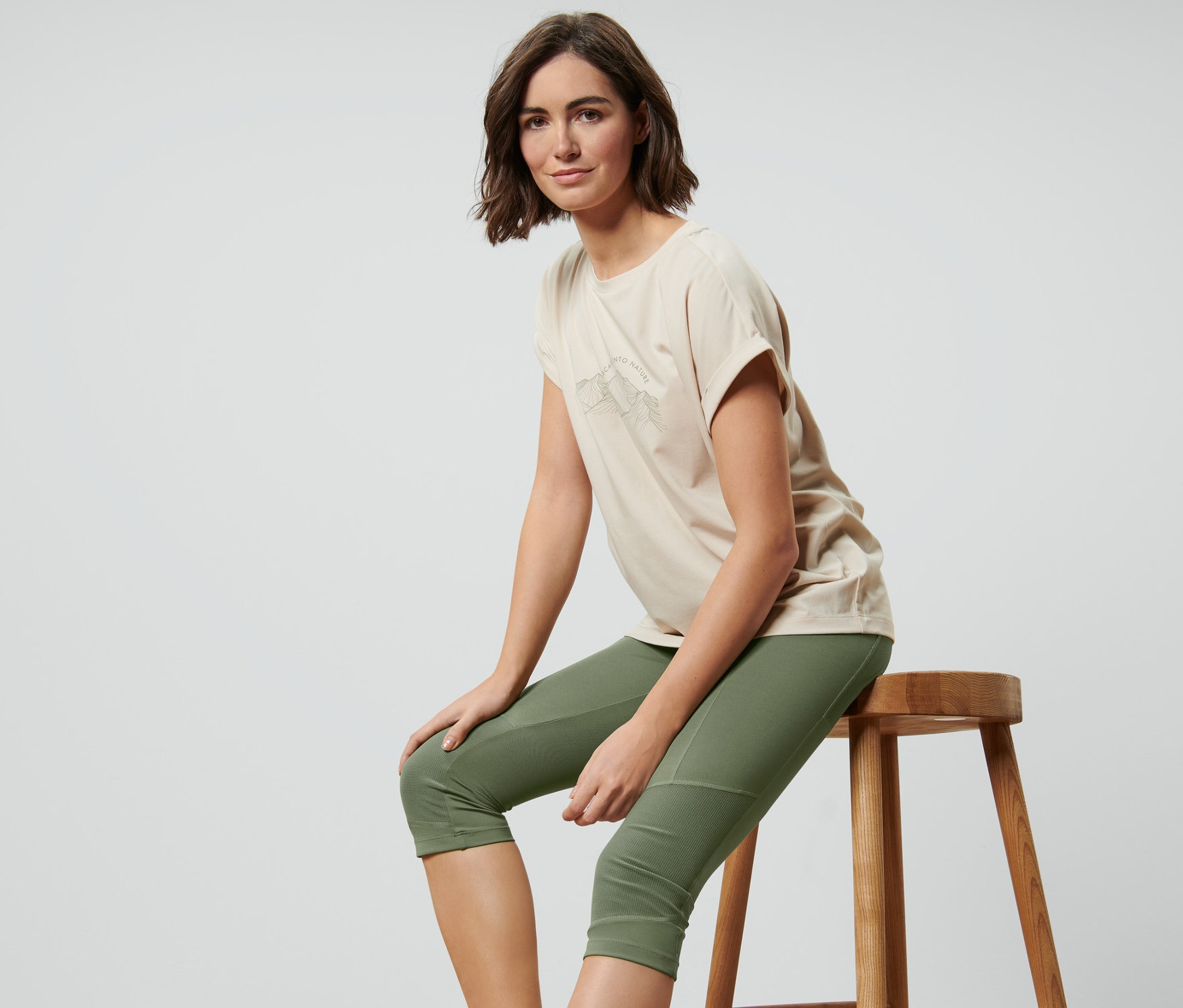 Kobieta siedzi na stołku, ubrana w legginsy trekkingowe o długości 3/4, oliwkowe.