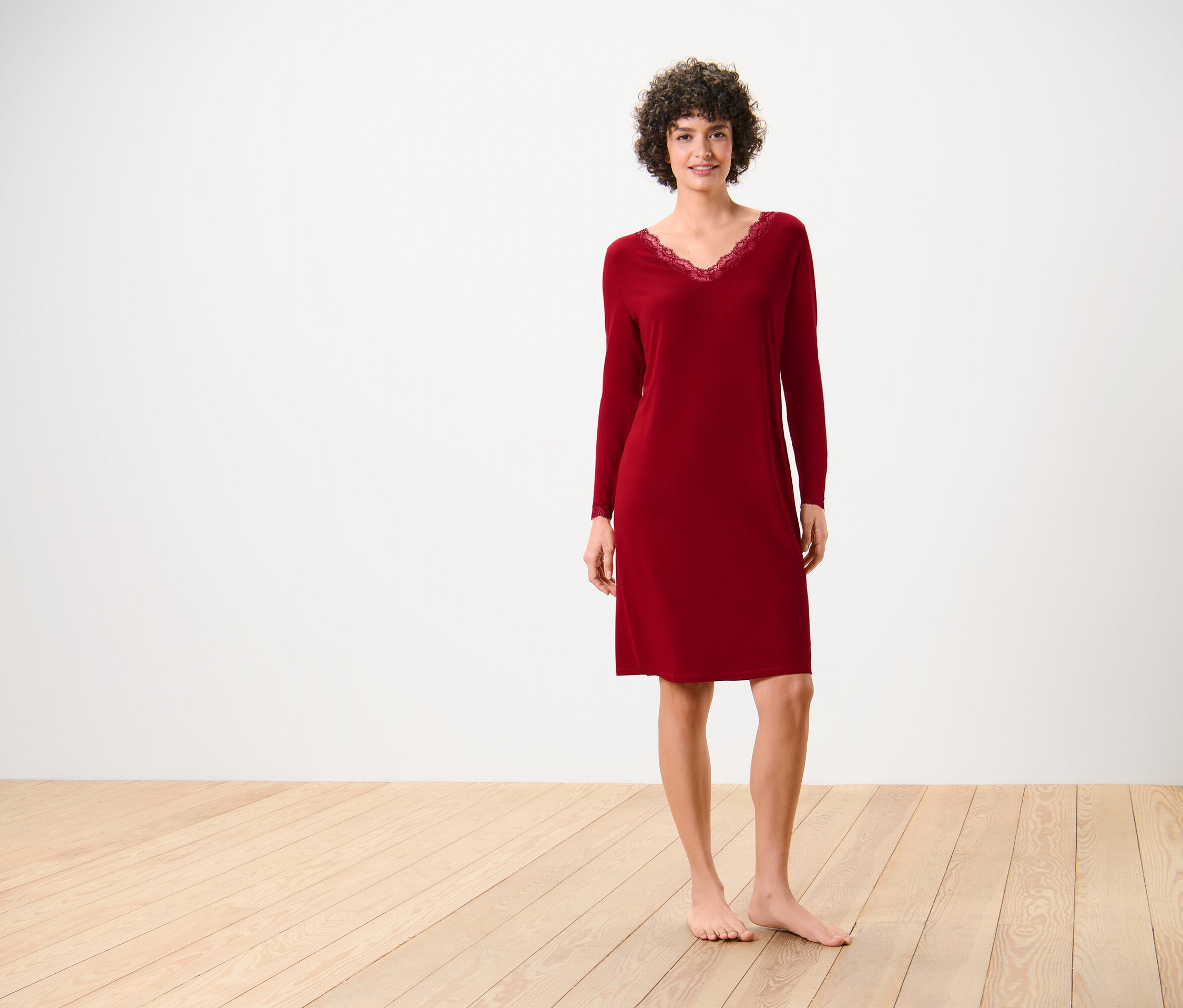 Kobieta w czerwonej koszuli nocnej z długim rękawem i koronką przy dekolcie stoi na drewnianej podłodze przed białą ścianą.