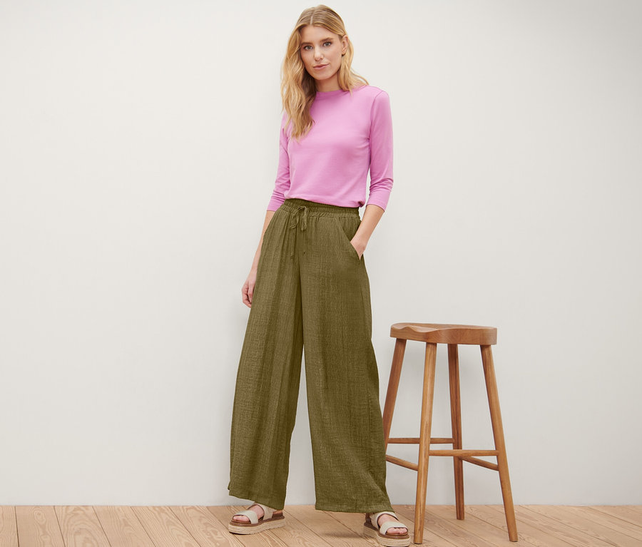 Kobieta pozuje w oliwkowych spodniach na gumce i różowym topie obok drewnianego stołka.