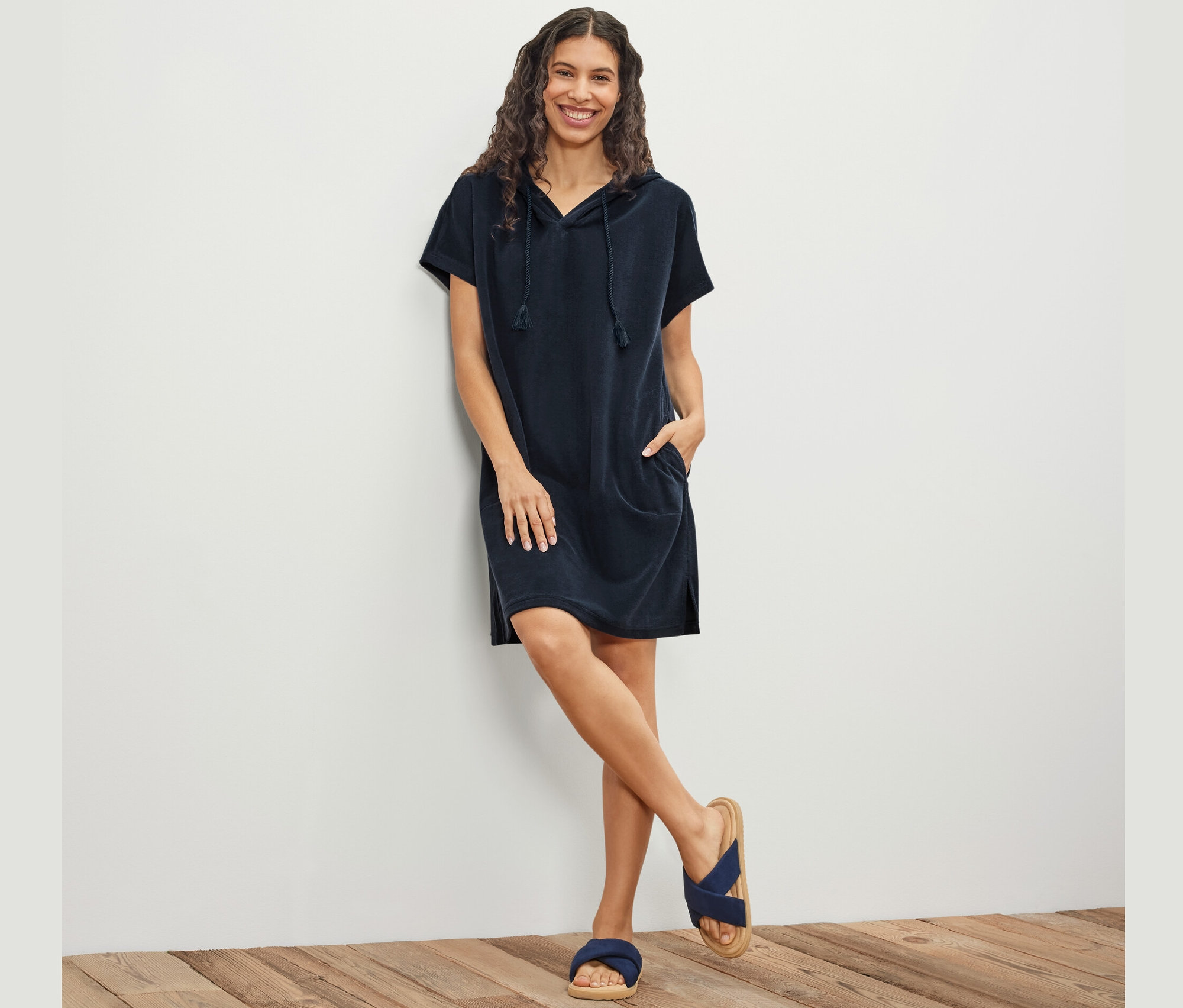 Kobieta pozuje oparta o ścianę, ubrana w niebieską sukienkę plażową z tkaniny frotte.