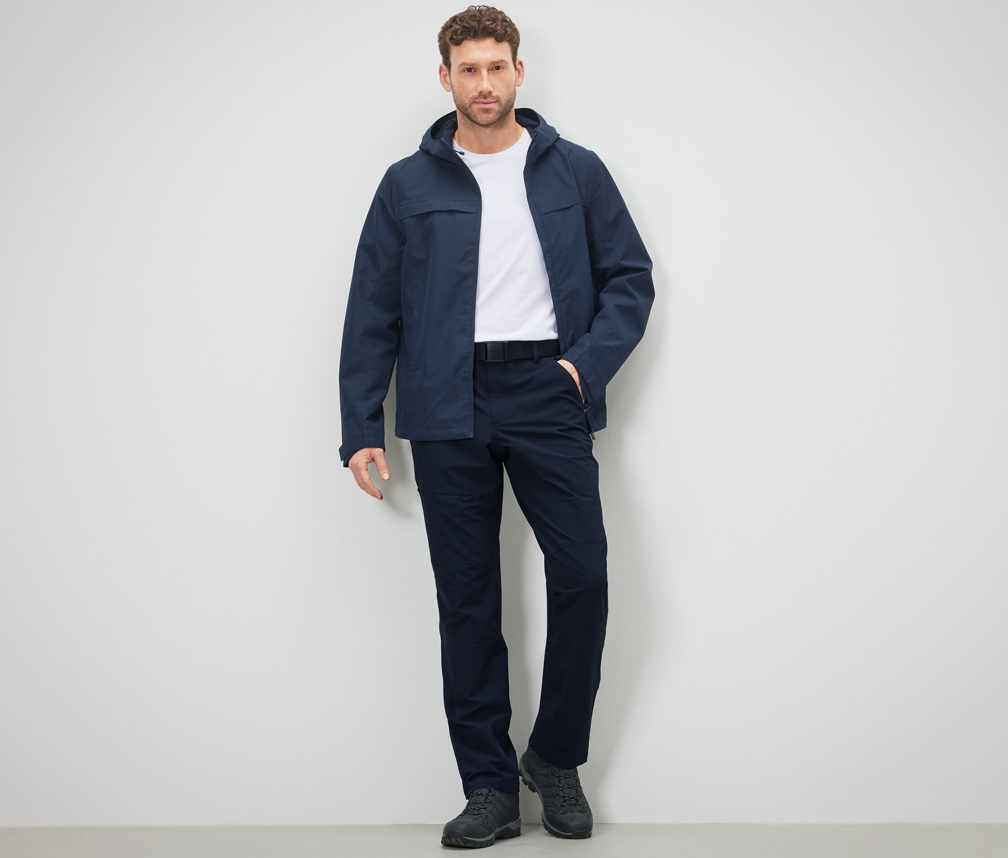 Mężczyzna pozuje w spodniach i kurtce outdoorowej na tle ściany.