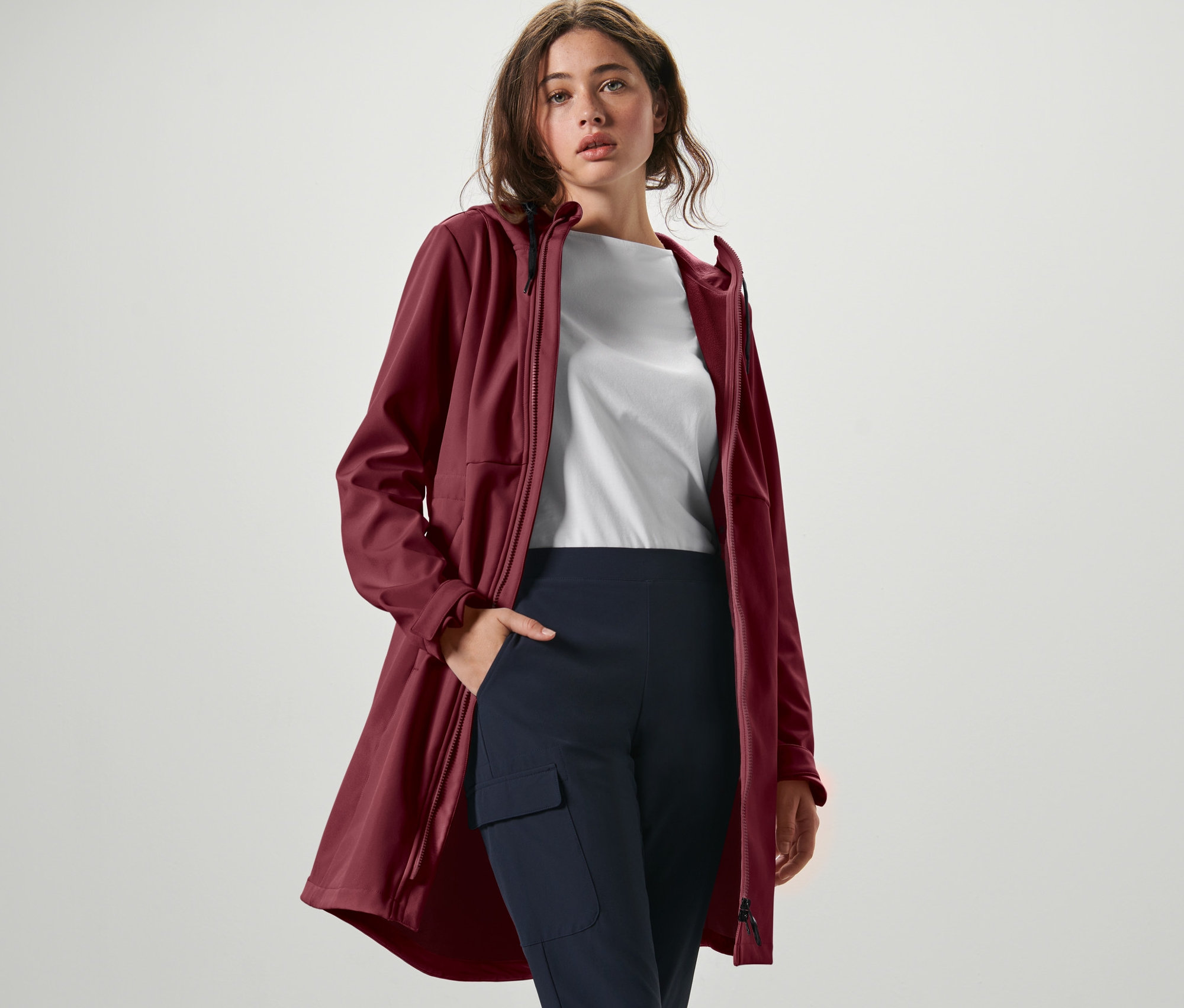 Kobieta pozuje w bordowym płaszczu softshell, białym T-shircie i granatowych spodniach.