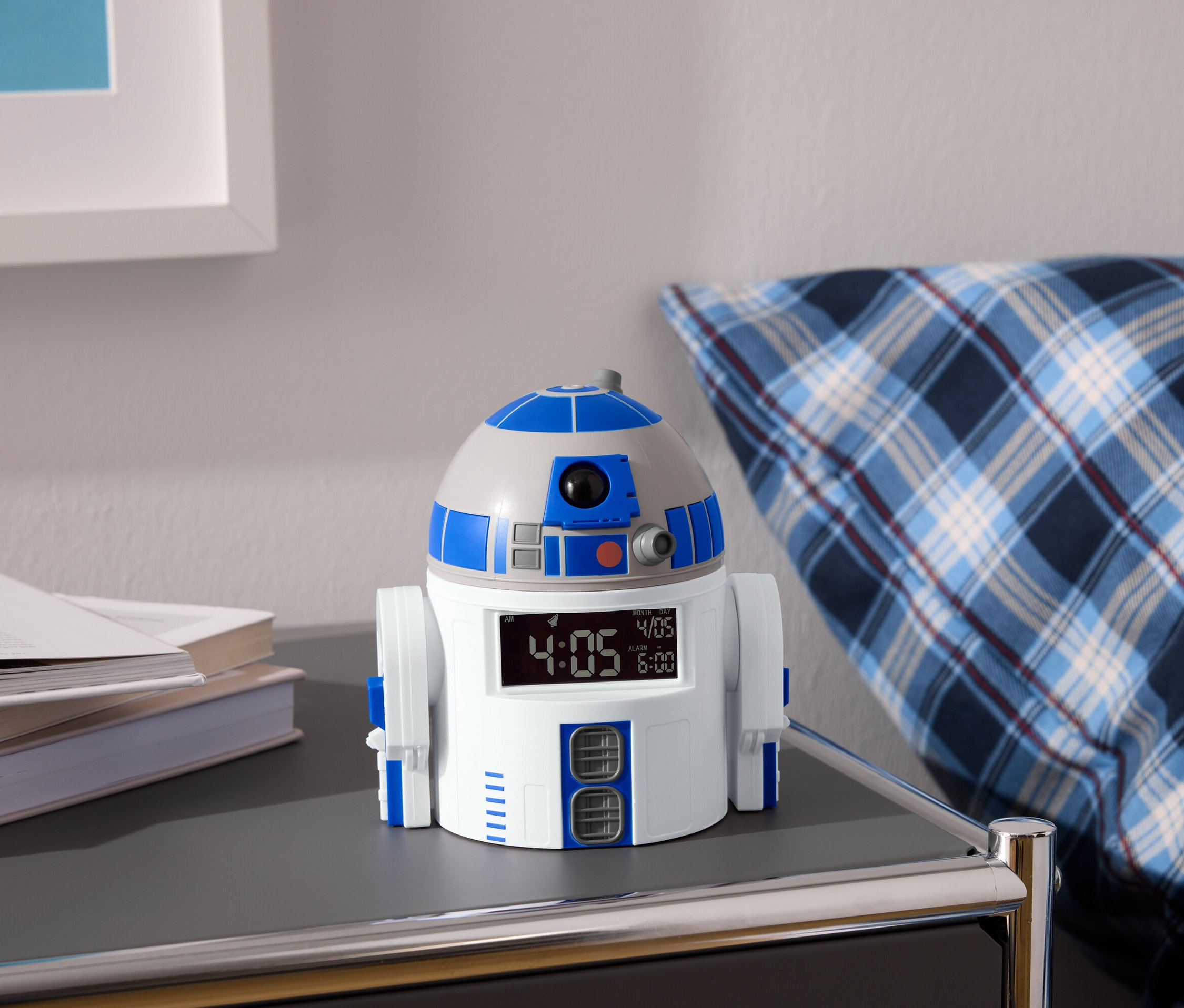 Budzik R2-D2 stoi na stoliku nocnym obok książek i niebiesko-białej poduszki w kratę.