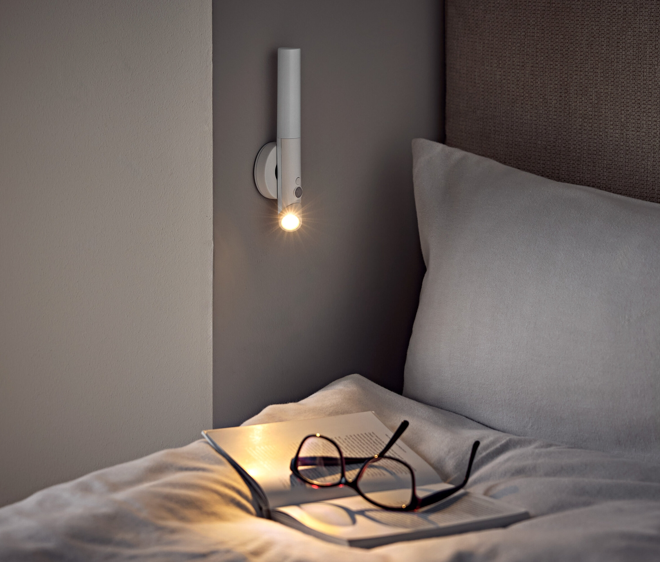 Na łóżku leżą okulary i otwarta książka pod lampą ścienną.