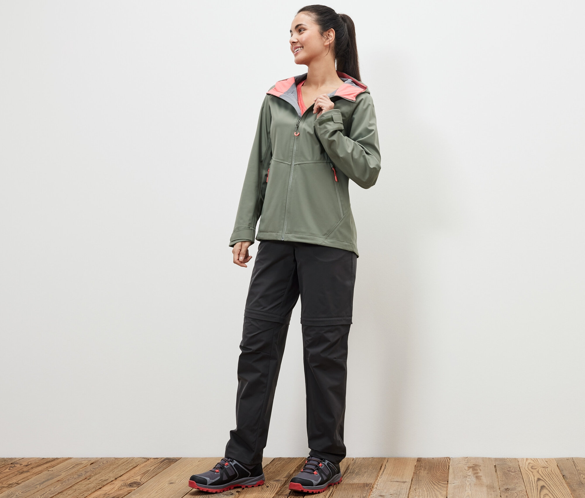 Kobieta stojąca na drewnianej podłodze ma na sobie zieloną kurtkę i czarne spodnie funkcyjne 2 w 1.