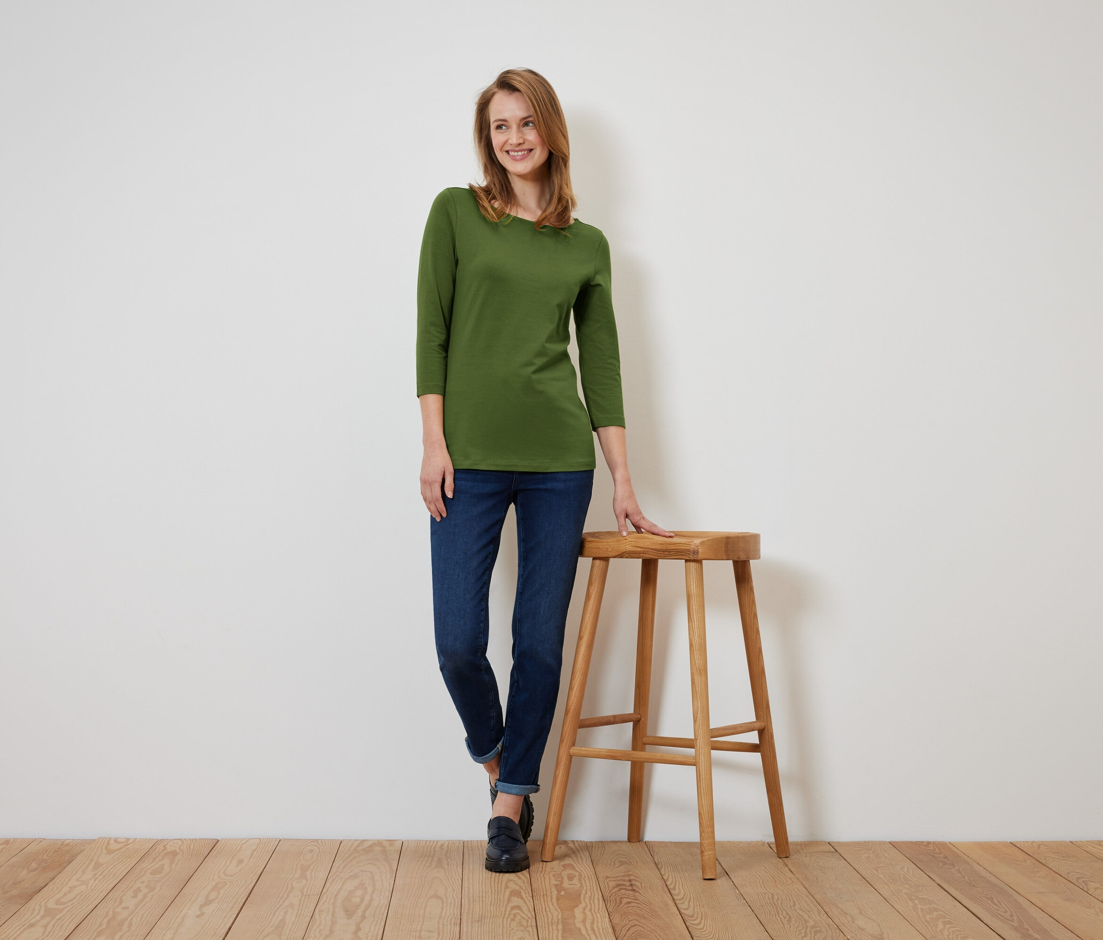 Kobieta w oliwkowozielonej koszulce z rękawem trzy czwarte i ciemnych dżinsach stoi, opierając się o drewniany stołek.