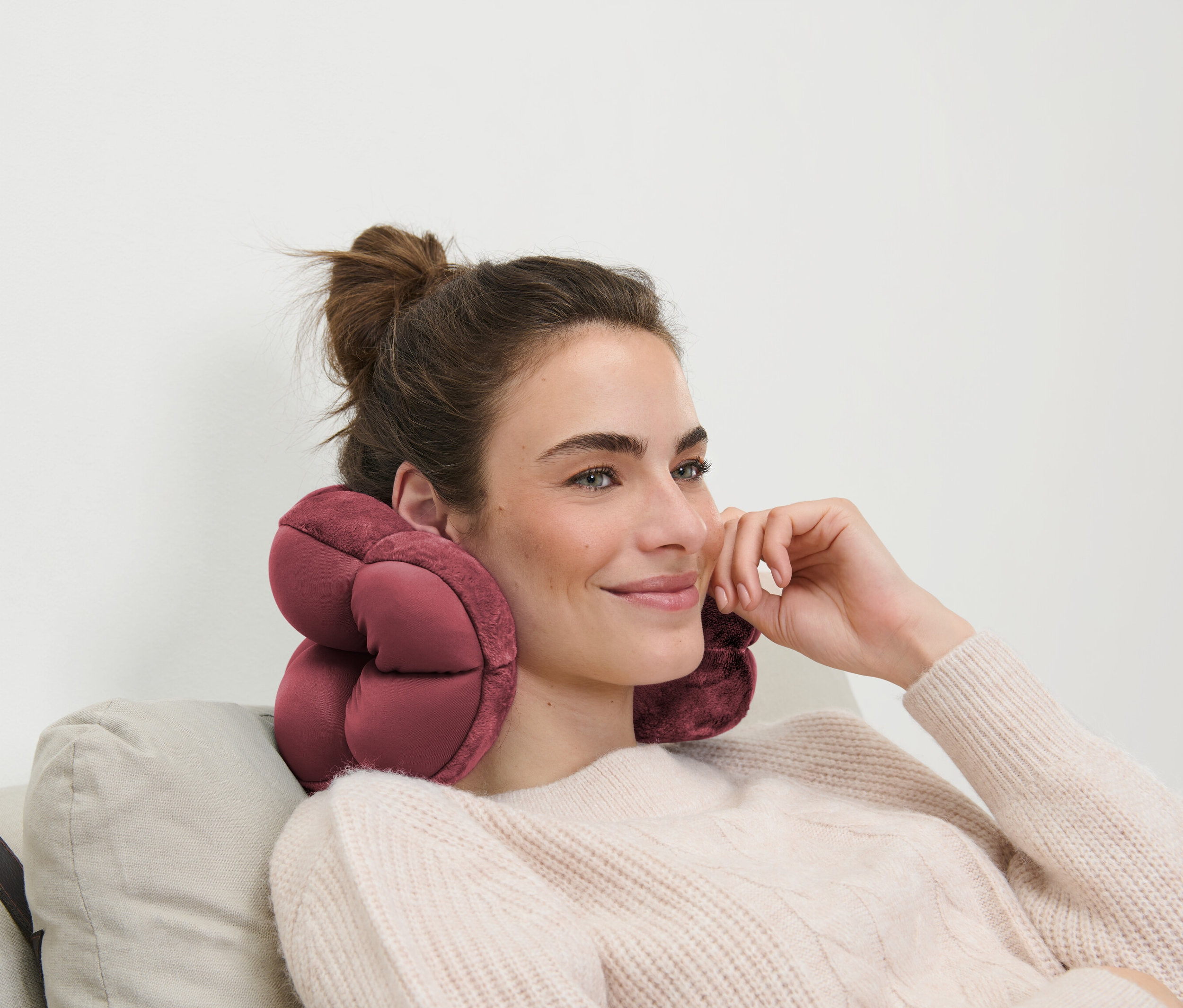 Kobieta z brązowymi włosami siedzi na sofie z różową poduszką podróżną na szyi.