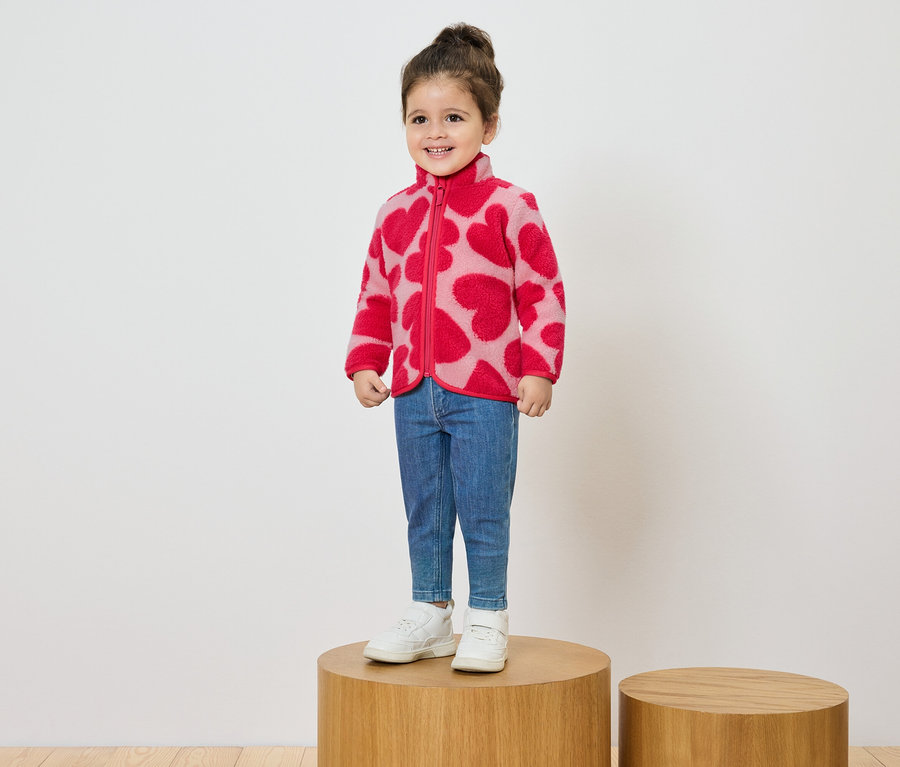 Uśmiechnięta dziewczynka stoi na drewnianym cokole, ubrana w różową kurtkę polarową w czerwone serca, dżinsy i białe tenisówki.