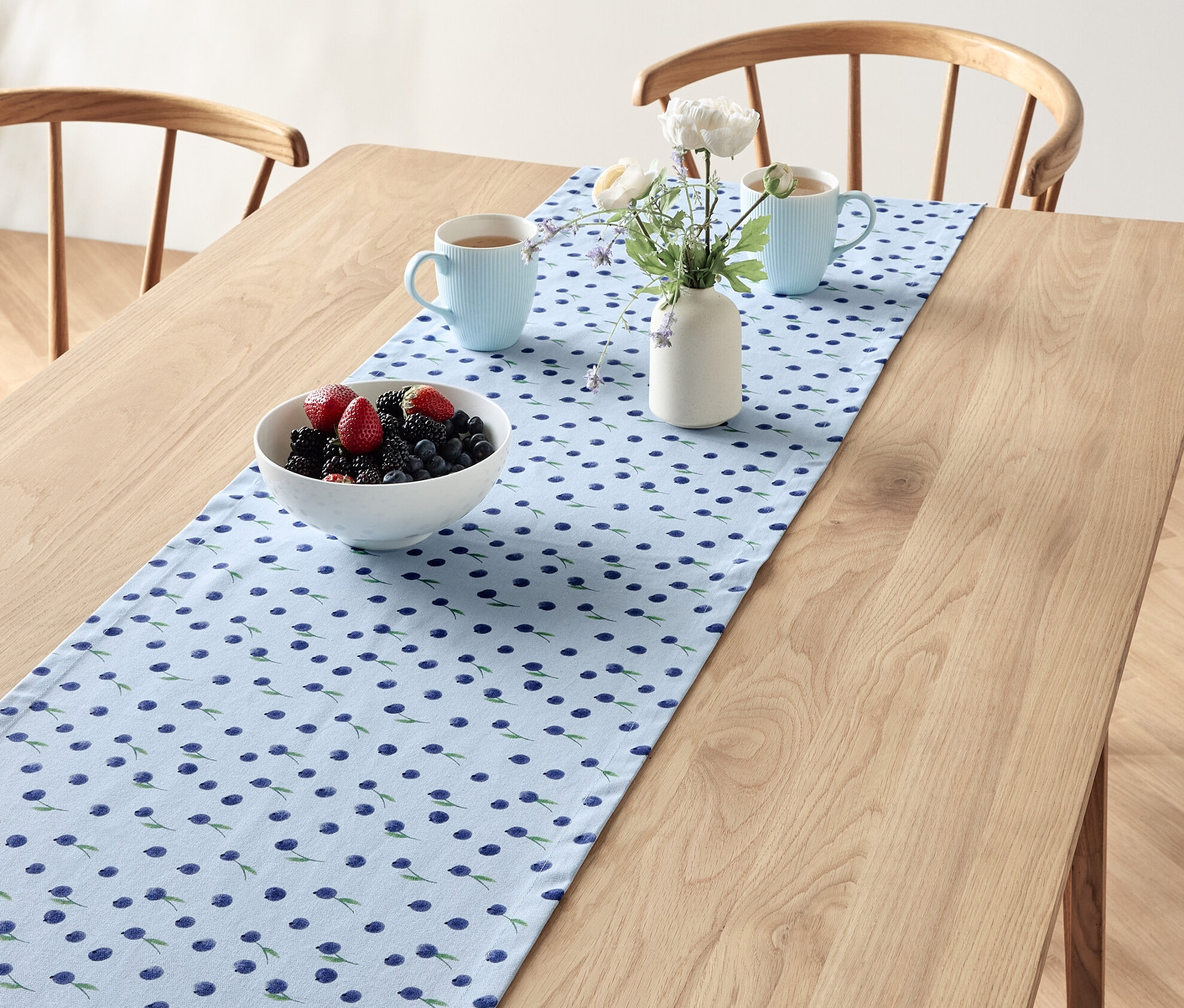 Na drewnianym stole leży bieżnik w borówki, na nim misa z owocami, dwa niebieskie kubki i wazon z kwiatami.