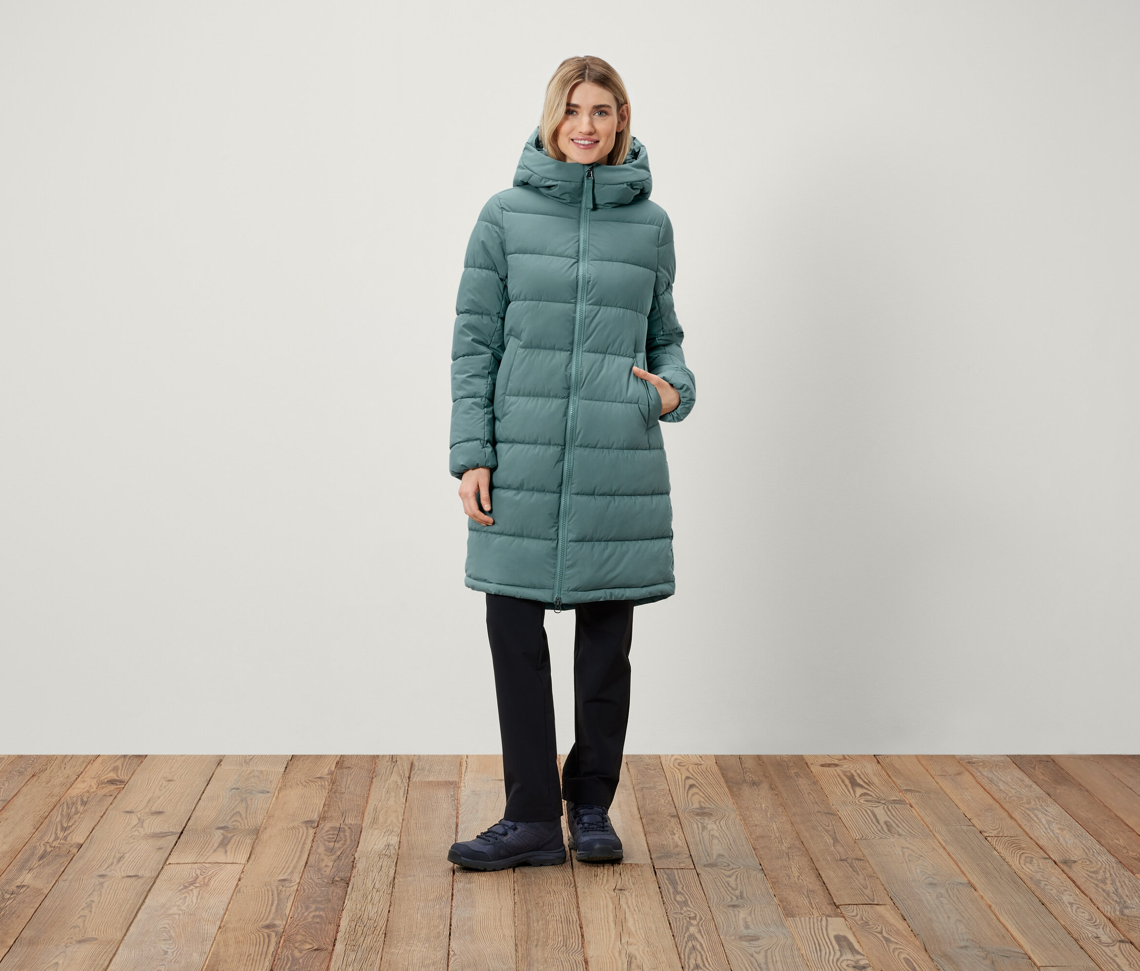 Kobieta stoi, ubrana w długi pikowany płaszcz z kapturem, czarne spodnie i buty.